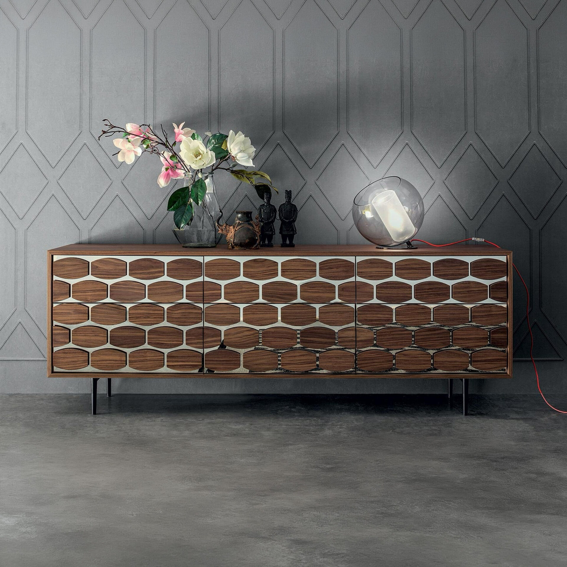 A modern wooden sideboard with a unique hexagonal and oval pattern, adorned with flowers, decorative figurines, and a spherical lamp, set against a dark gray geometric wall and concrete floor.