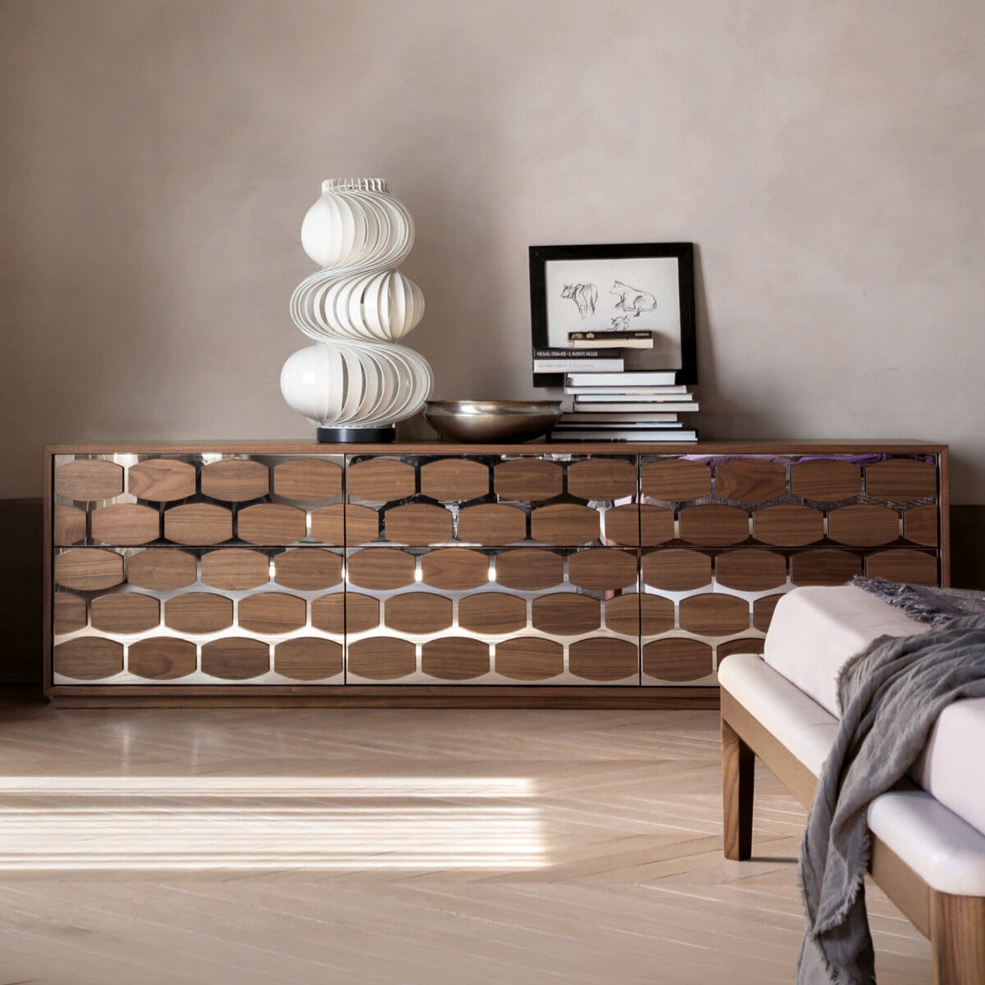 A modern credenza with dark wood and reflective hexagonal panels, topped with a sculptural white lamp, a metallic bowl, and a stack of books with a framed sketch. The room has light wood floors with sunlit shadows and a neutral wall.
