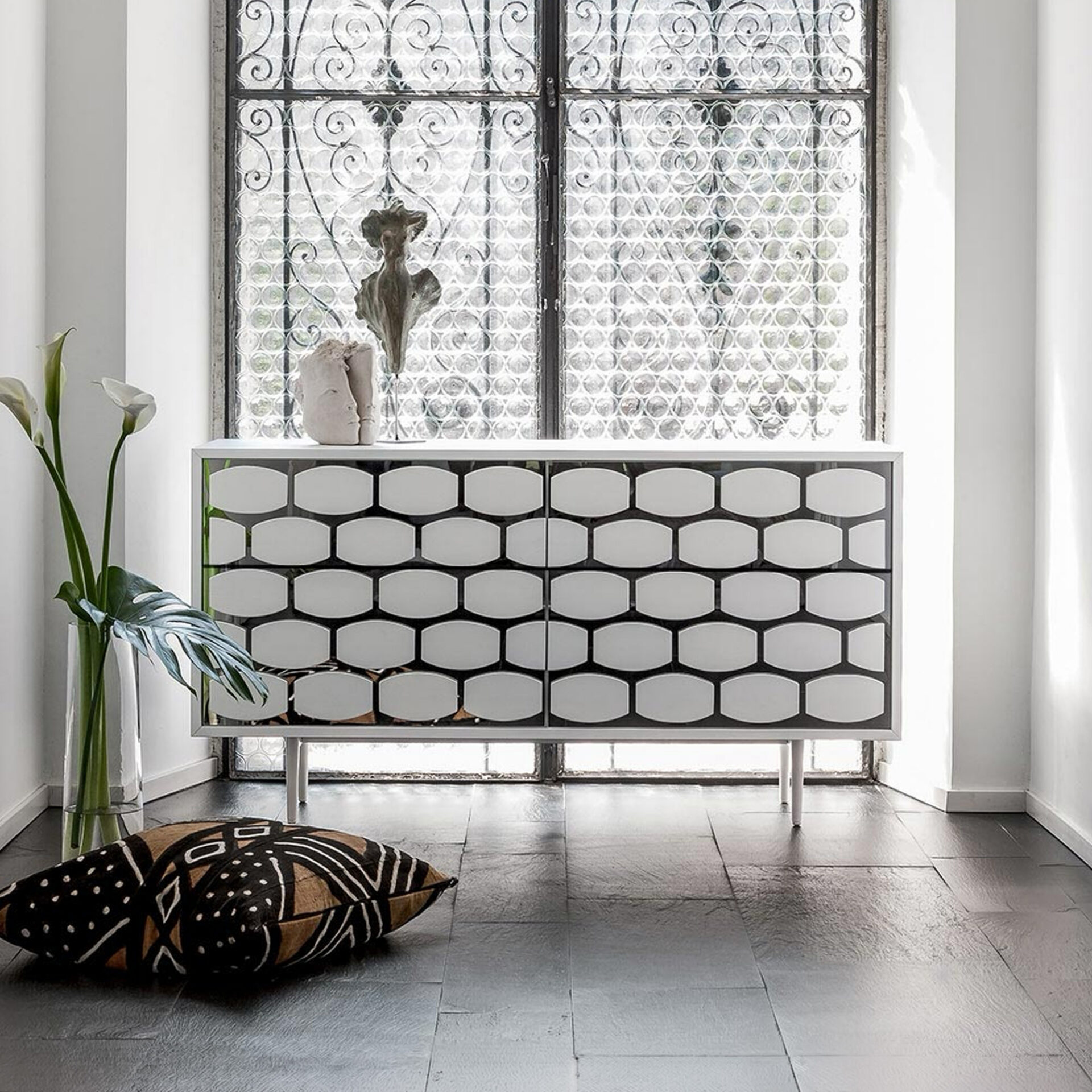 A modern white credenza with a geometric pattern on its front sits in front of a large window with textured glass and intricate wrought iron grilles. A vase with white calla lilies and a monstera leaf is to the left, and a patterned floor cushion rests on dark tiles in the foreground.