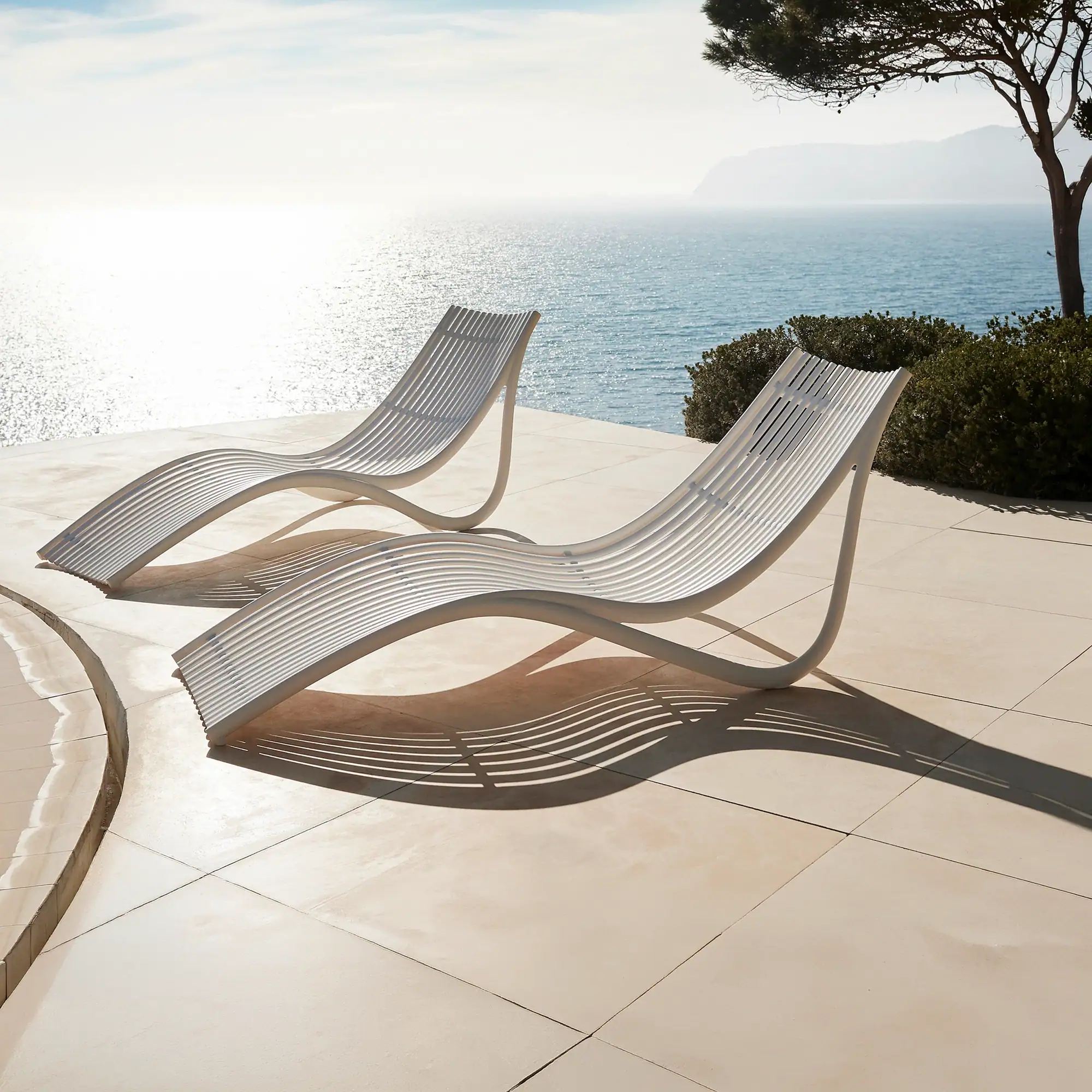 Two white, curved lounge chairs on a tiled patio overlooking a sparkling blue ocean under a bright sky.