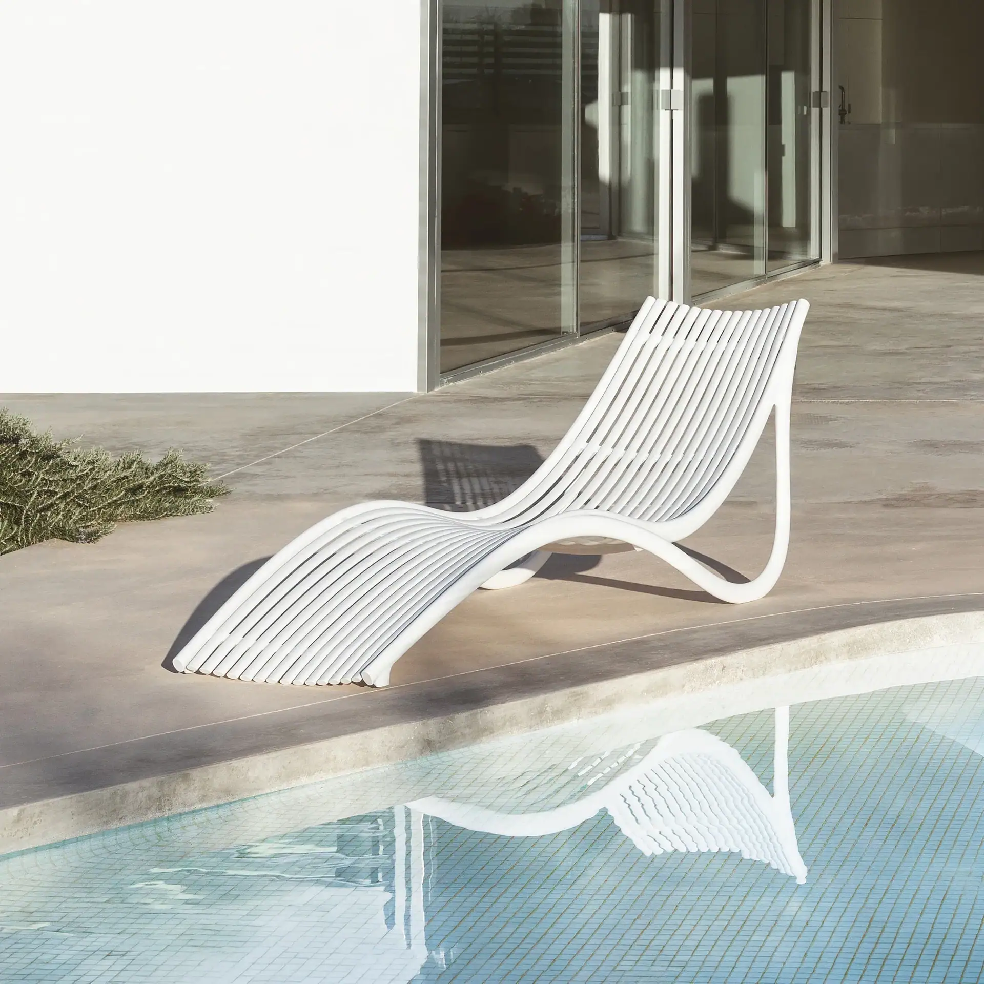 A white slatted lounge chair on a concrete patio next to a swimming pool, with a building and glass doors in the background.