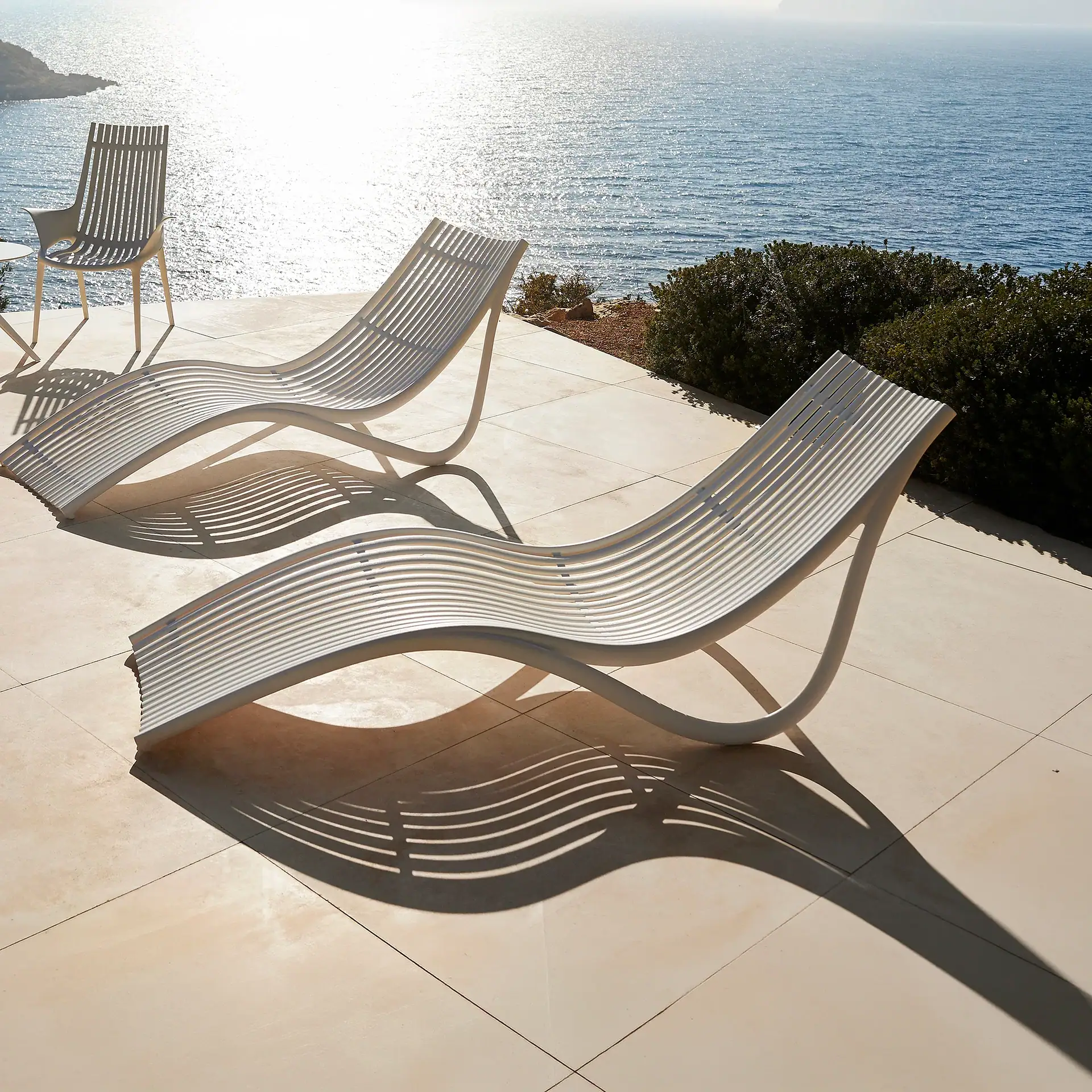 Two white slatted lounge chairs and a dining chair on a tiled patio overlooking a sparkling blue ocean under bright sunlight.