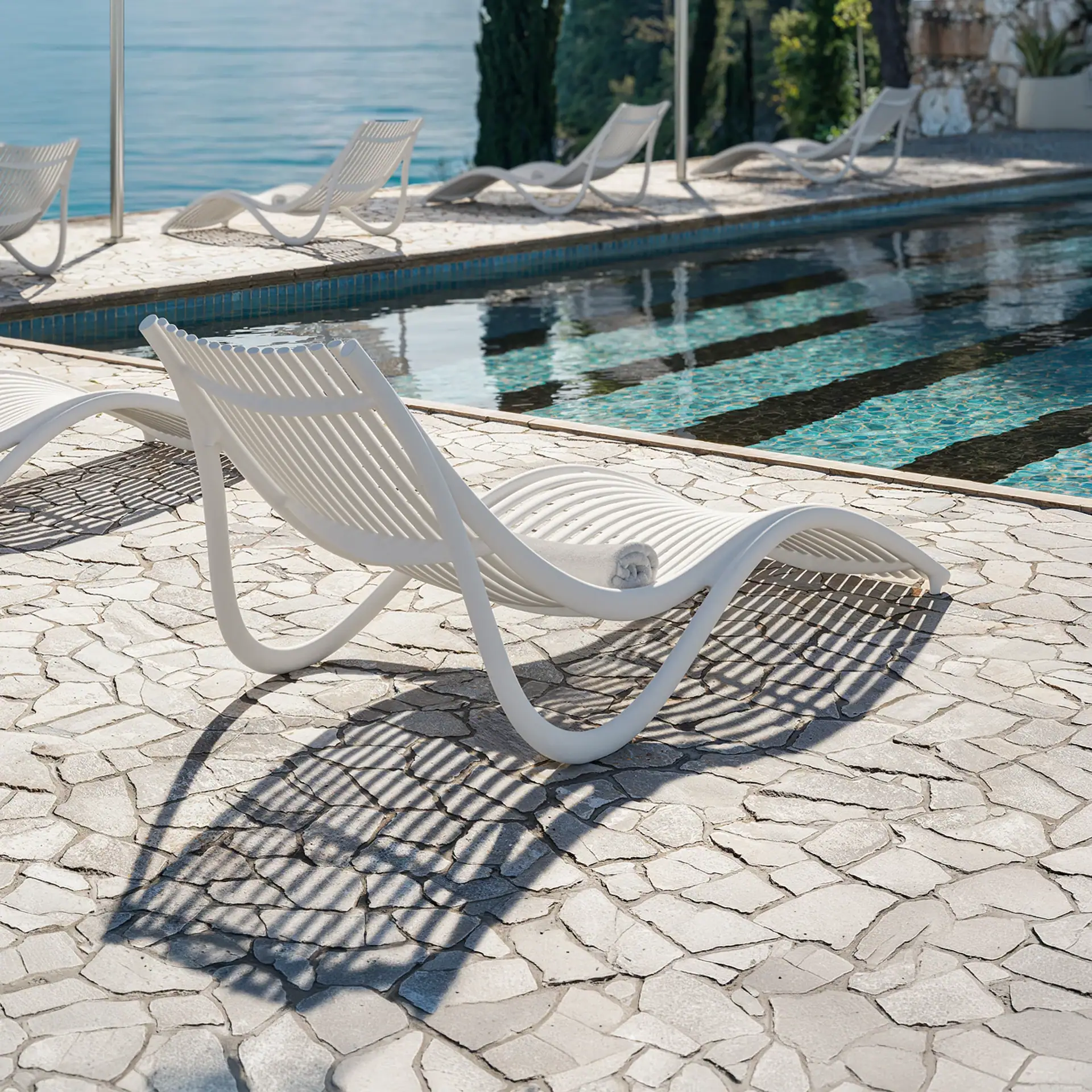 A white slatted lounge chair with a rolled towel on a stone patio next to a swimming pool, with more chairs, green trees, and blue water in the background.