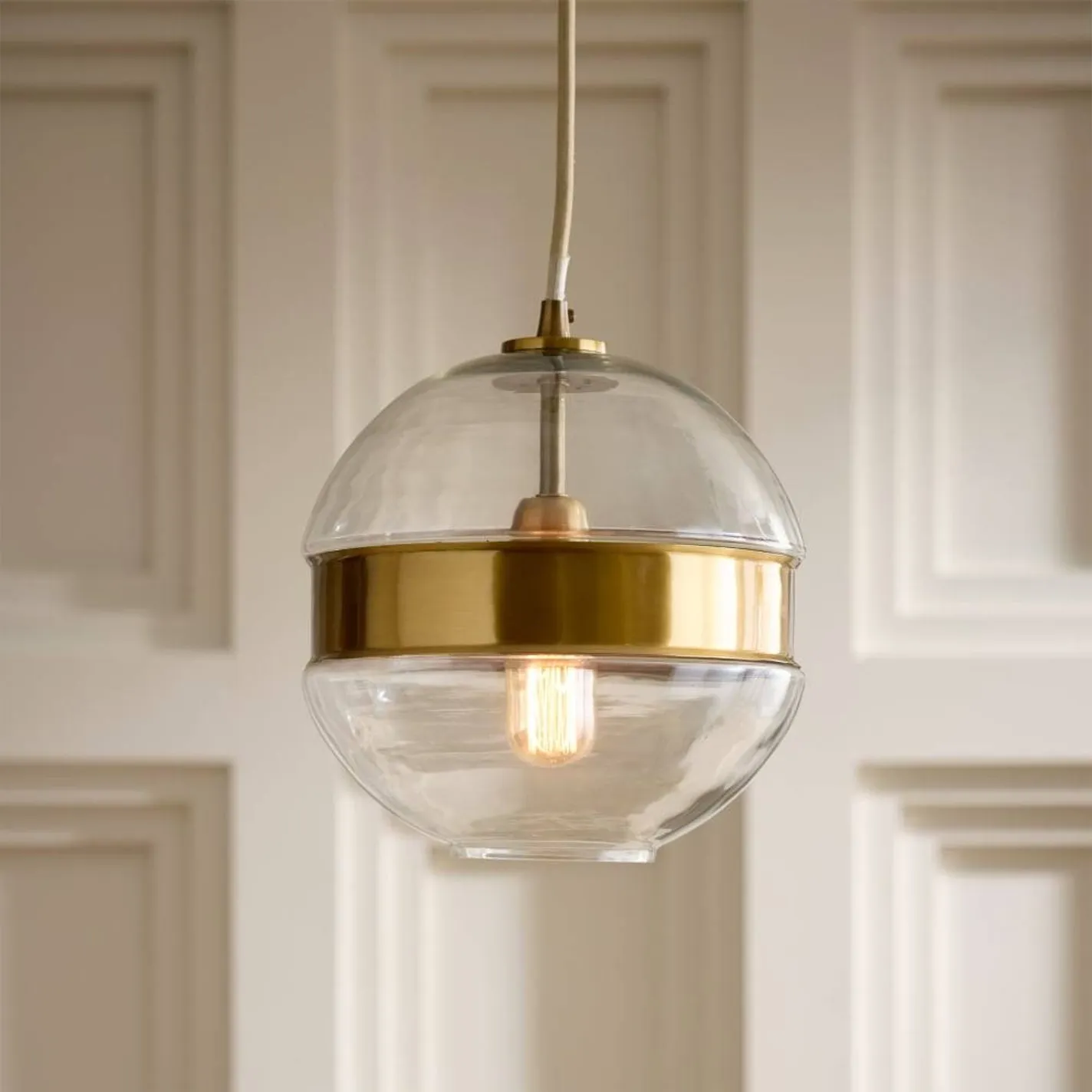 A modern globe pendant light with a clear glass shade and a brushed brass band, featuring a glowing Edison bulb, hanging against a light-colored paneled wall.