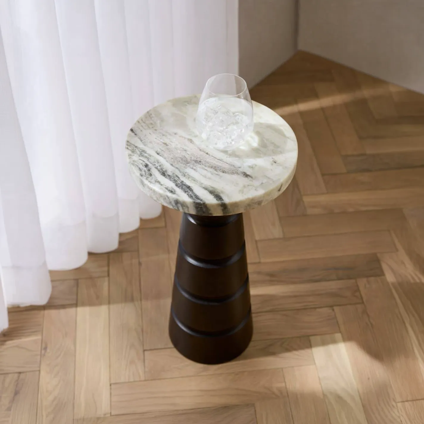 A modern side table with a round, veined marble top and a dark, tiered base. A glass of ice water sits on the table, next to sheer white curtains and a herringbone wood floor.