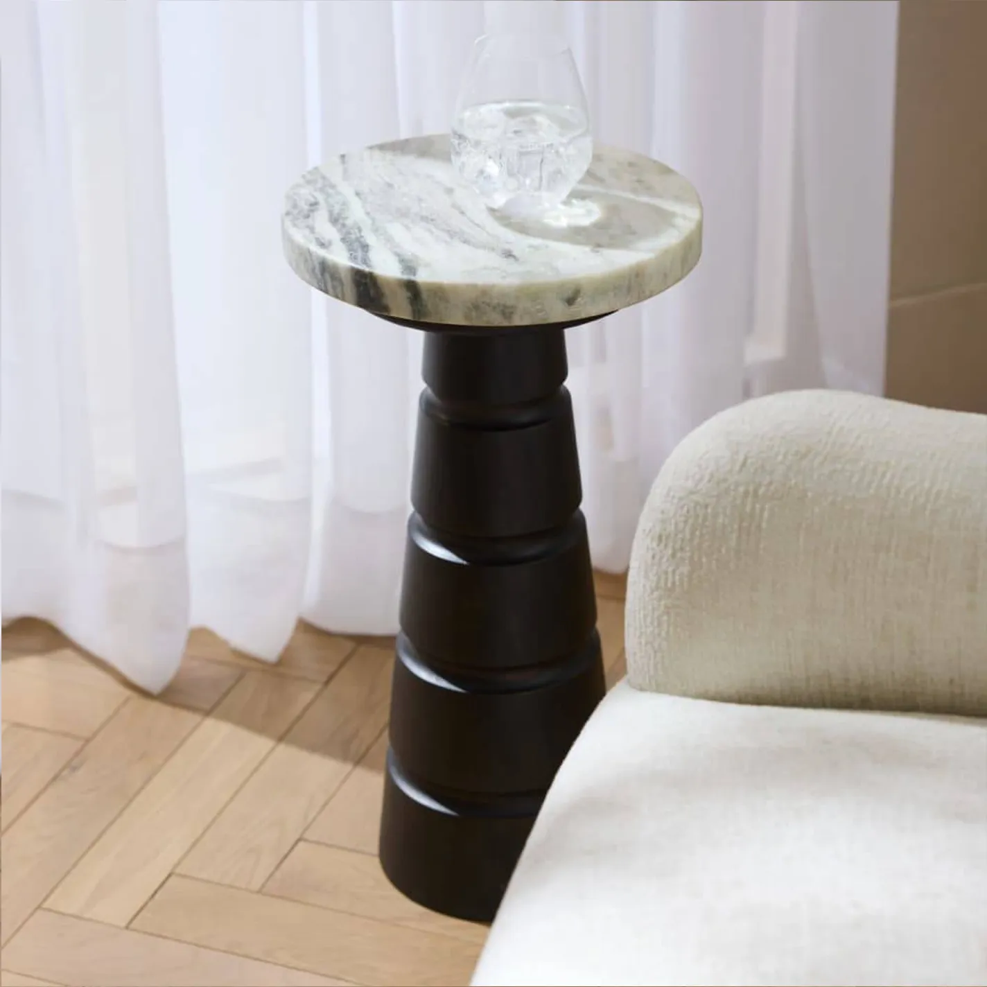A modern side table with a round marble top and a dark, sculptural tiered base, next to a cream upholstered chair. Sheer white curtains are in the background, and the floor is light wood in a herringbone pattern.