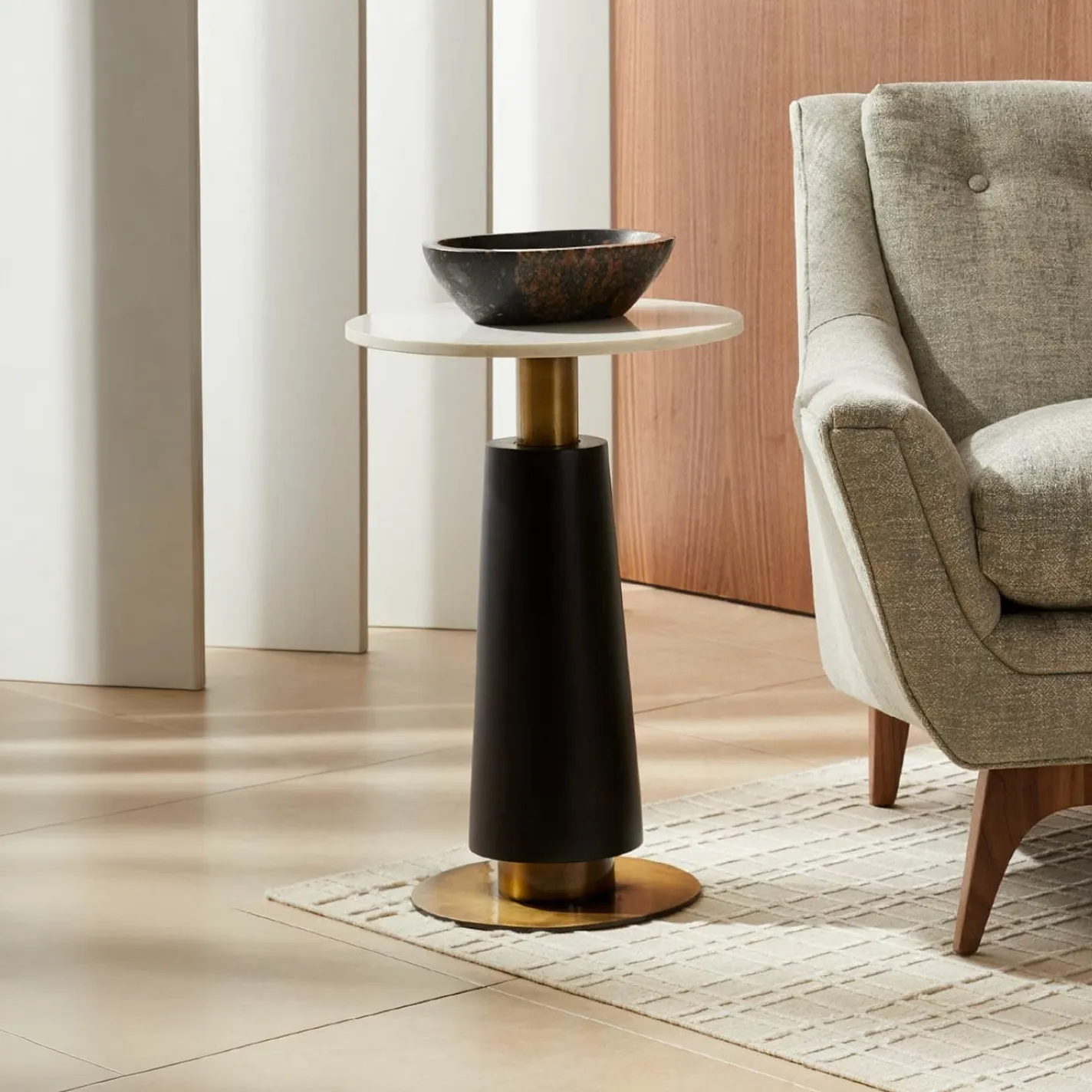 A modern side table with a white marble top, black tapered column, and brass accents, next to a gray fabric armchair. A dark decorative bowl is on the table.