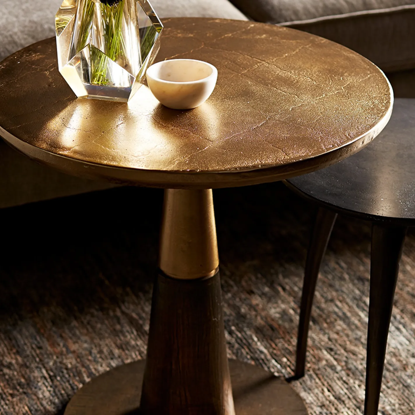 A round side table with a textured gold top, holding a faceted glass vase with greenery and a small light-colored bowl. A dark side table and a textured rug are visible in the background.