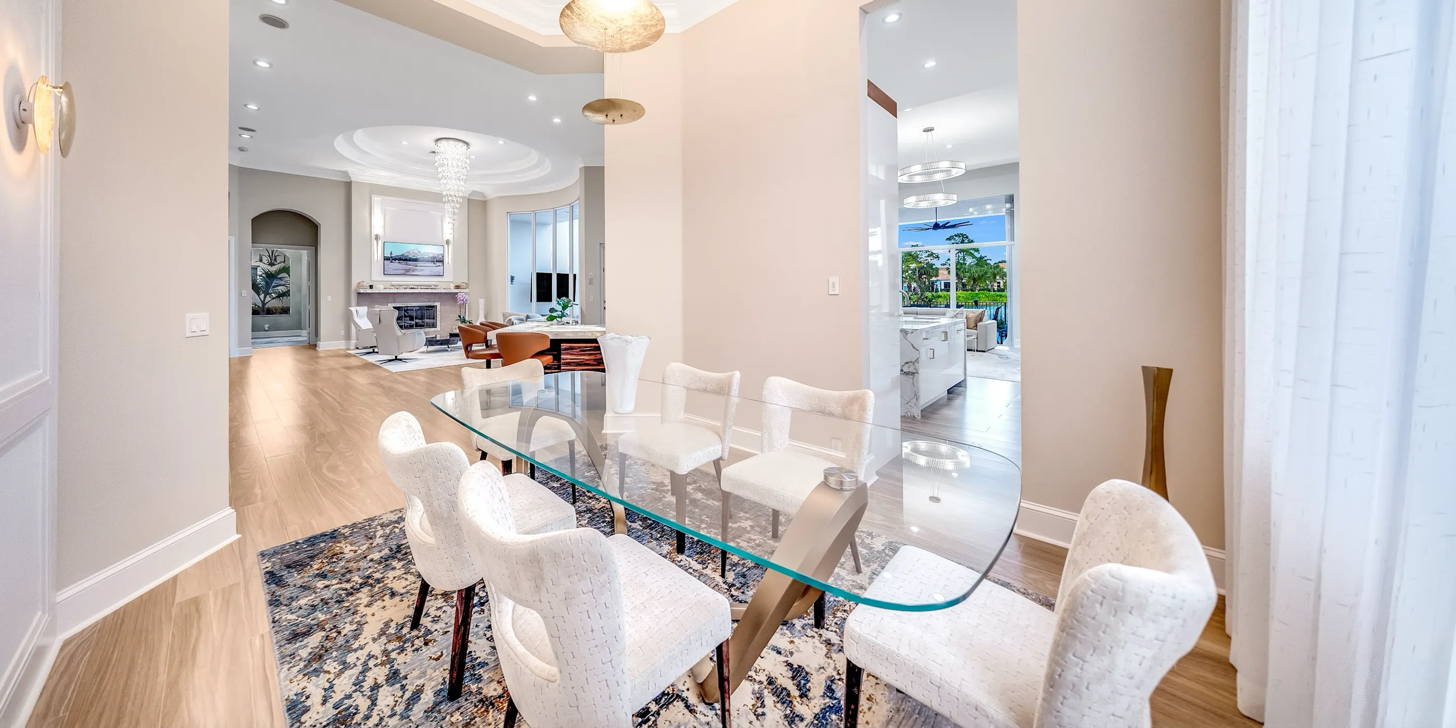 A modern open-concept dining room with a glass table and eight white upholstered chairs on an abstract rug. The room opens to a living room with a fireplace and a kitchen with water views.