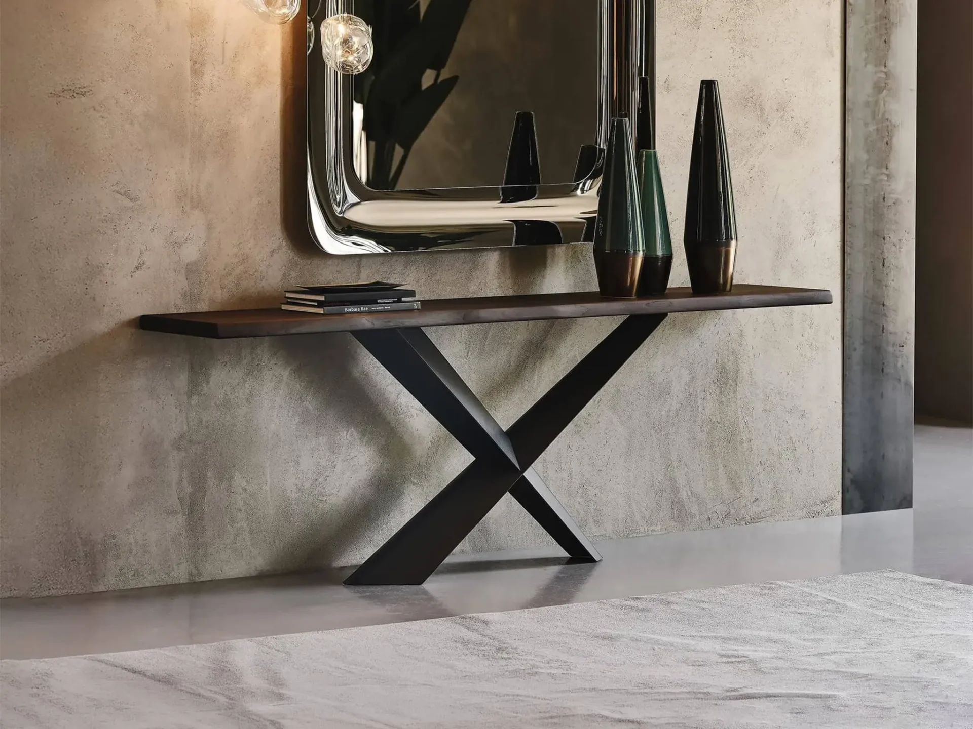 A modern console table with a dark wood top and black X-shaped base, with a large silver-framed mirror above it. On the table are books and three decorative vases.
