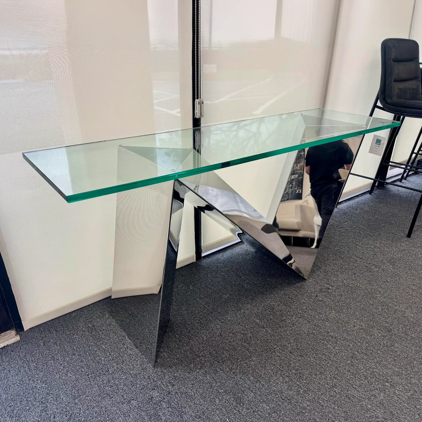 A modern console table with a clear glass top and a geometric, mirrored base, reflecting its surroundings.