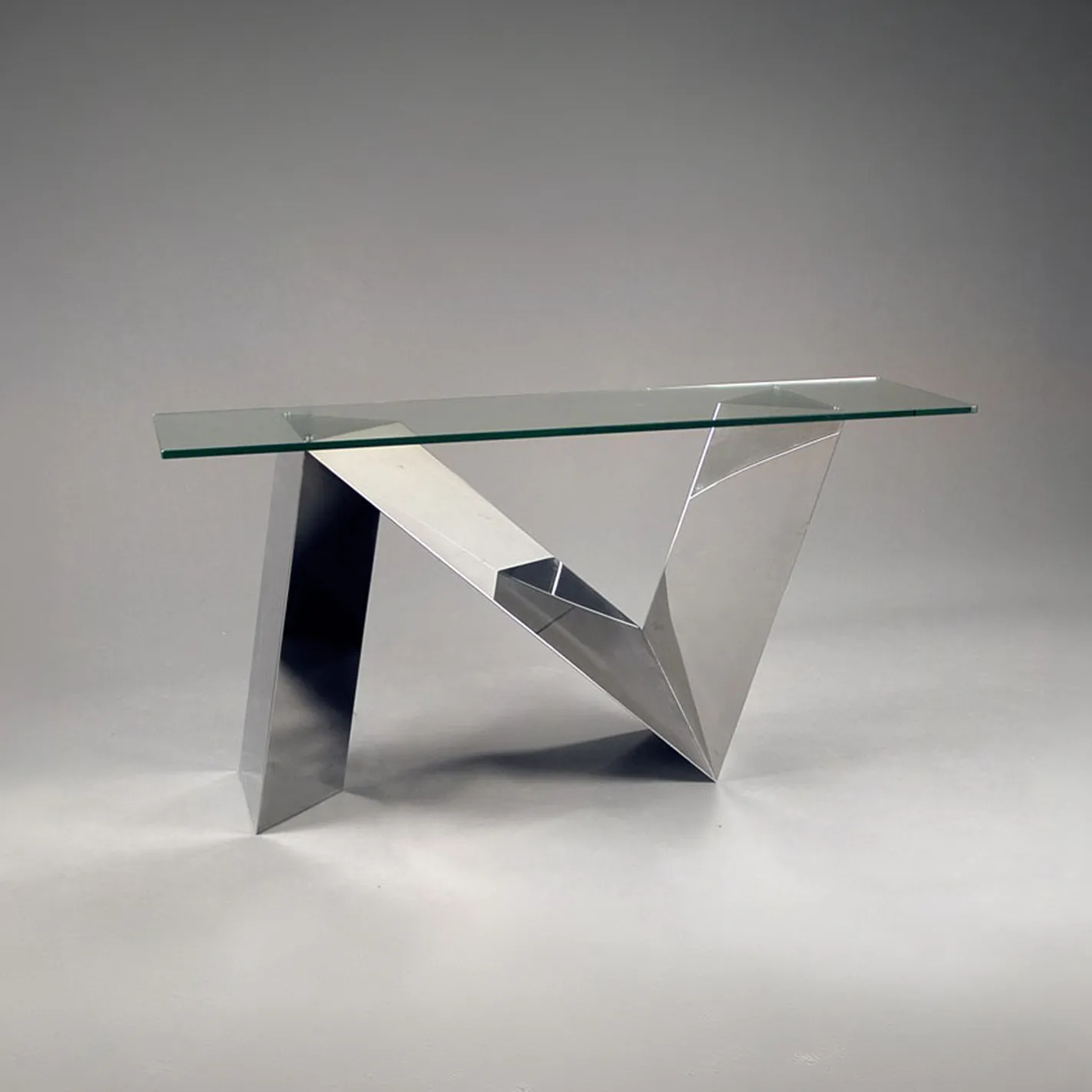A modern console table with a clear glass top and a highly polished, faceted metal base in a sculptural, geometric design, set against a gray background.