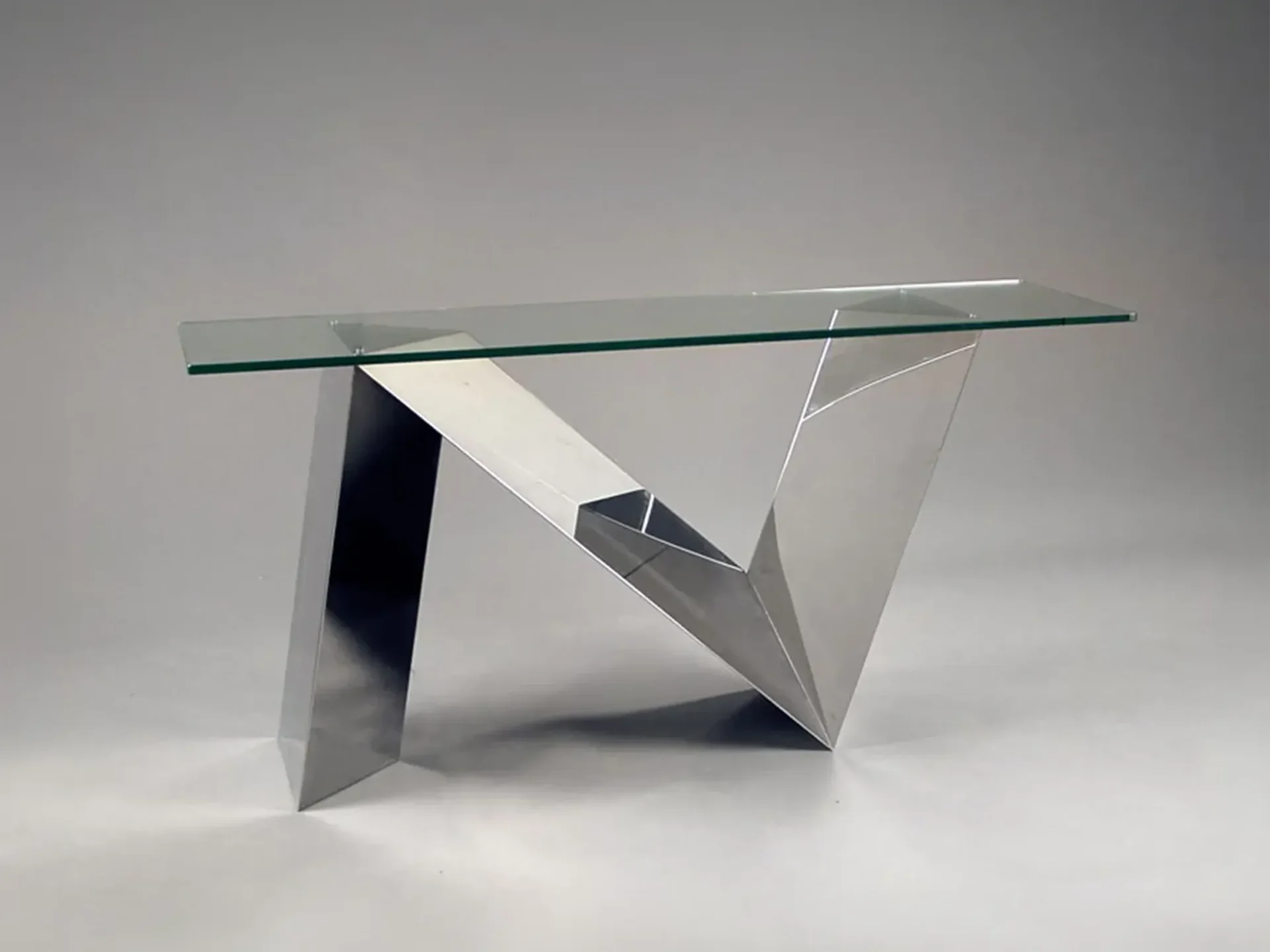 A modern console table with a clear glass top and a highly geometric, polished silver metal base against a gray background.