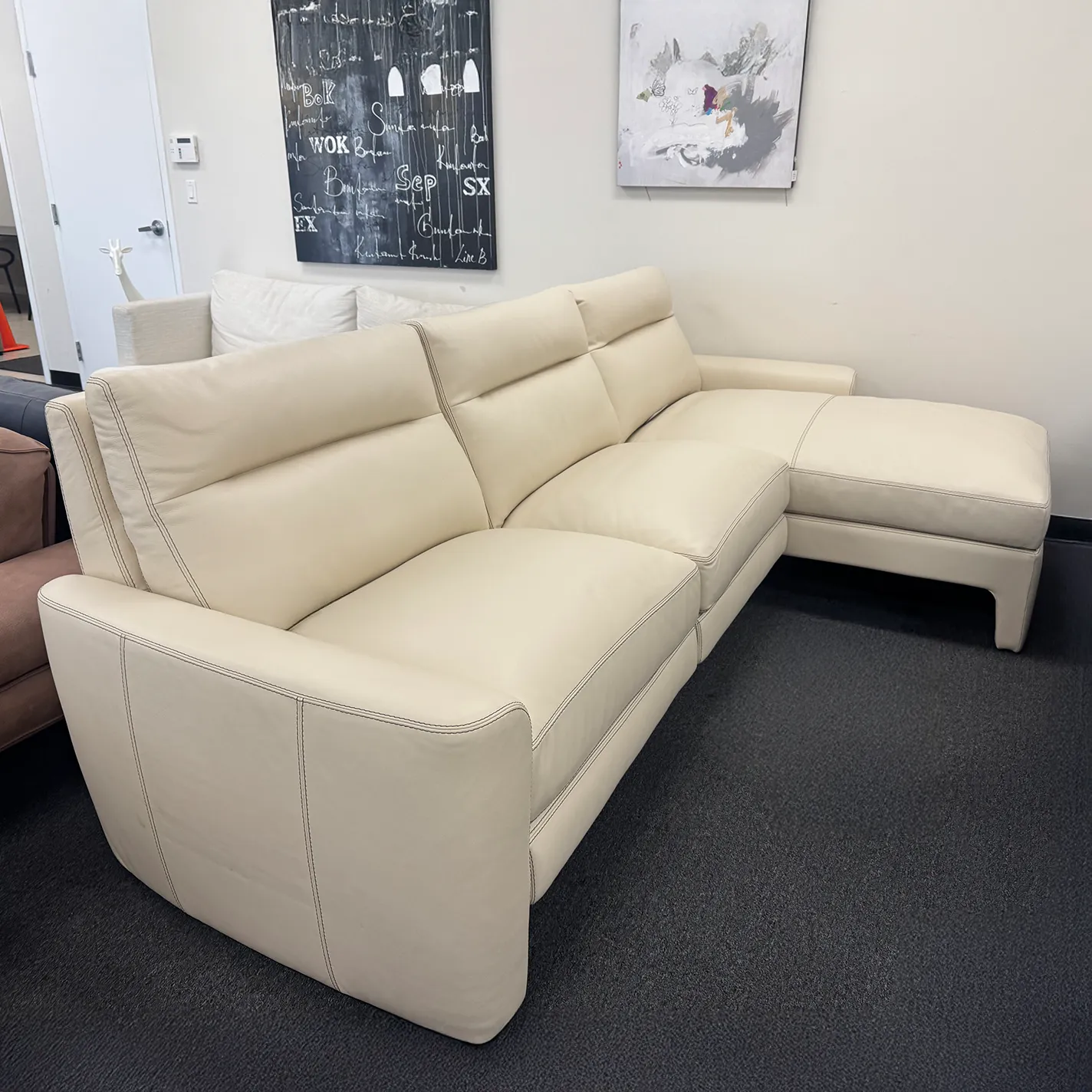 A cream-colored L-shaped sectional sofa with a right-hand chaise, dark stitching, and adjustable headrests, set on a dark gray carpet in a showroom with abstract wall art.