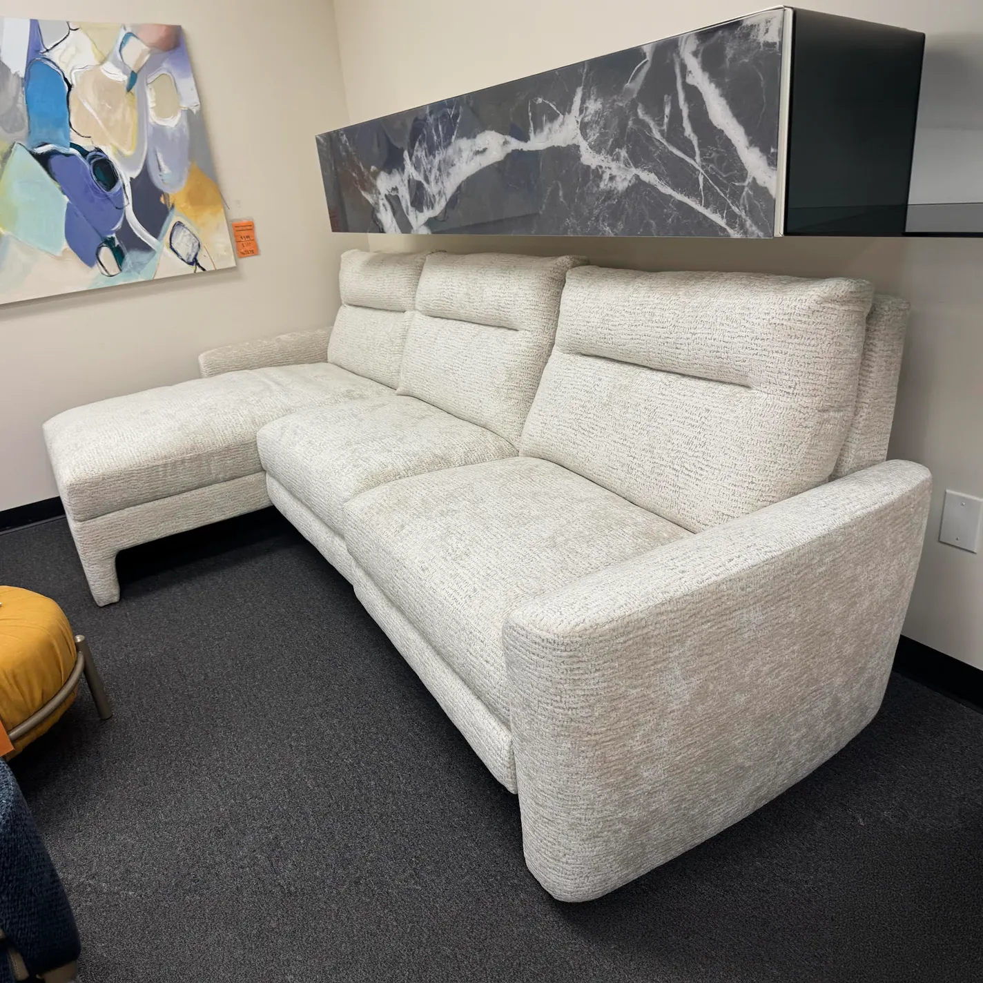 A light beige L-shaped sectional sofa with a chaise lounge on a dark gray carpet. Above it, a wall-mounted unit with a marble pattern and an abstract painting are visible.