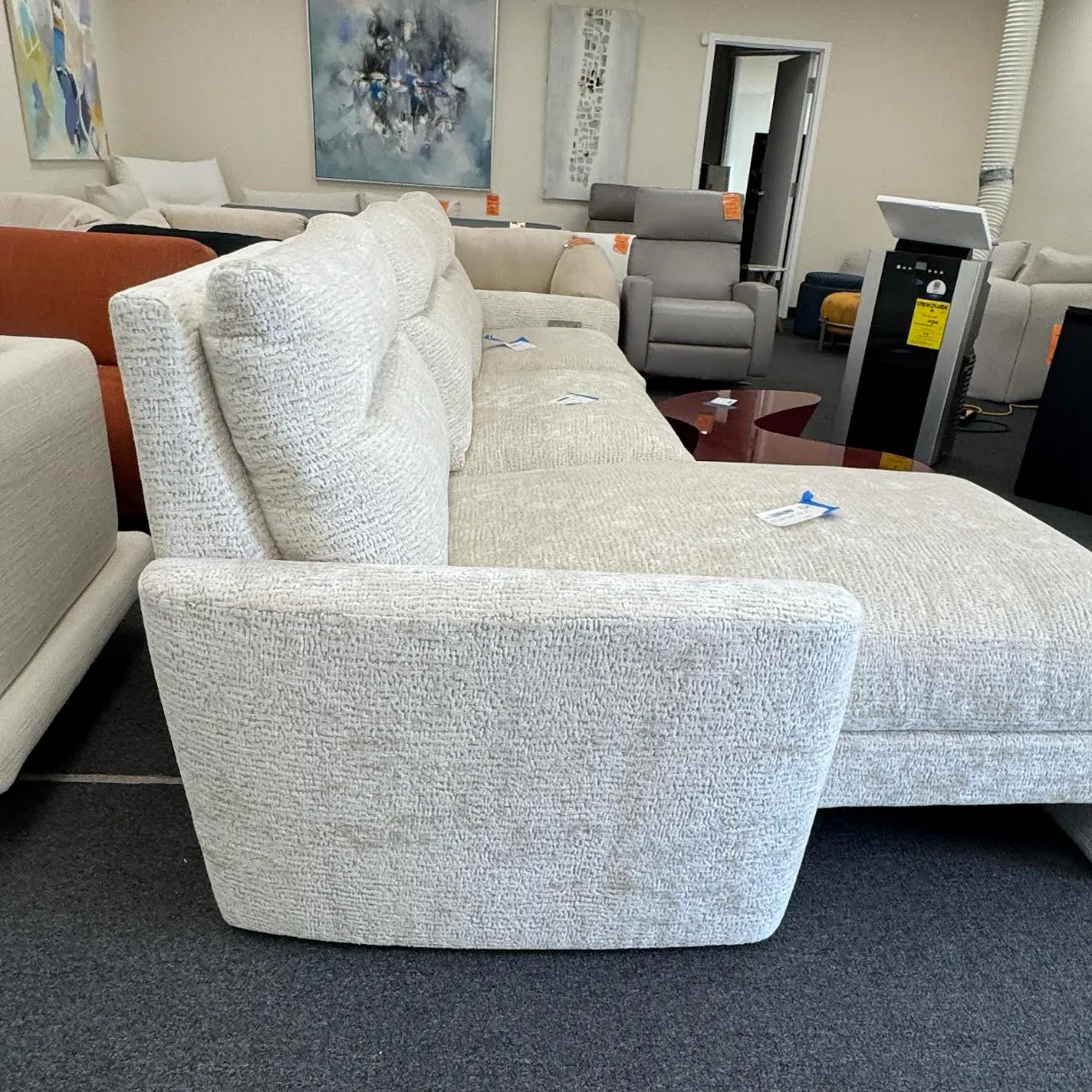 A light gray textured sectional sofa with curved armrests in a furniture showroom, surrounded by other modern furniture and abstract art.