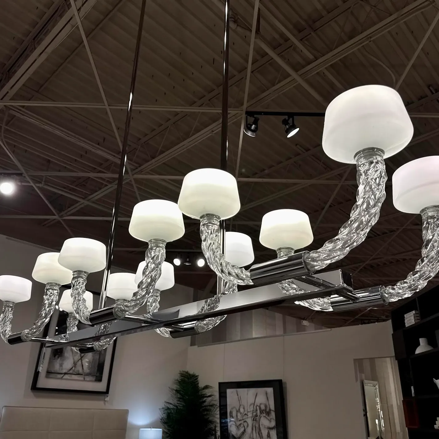 A modern linear chandelier with ten frosted white glass shades and twisted clear glass arms, suspended from an industrial ceiling in a room with artwork and furniture.