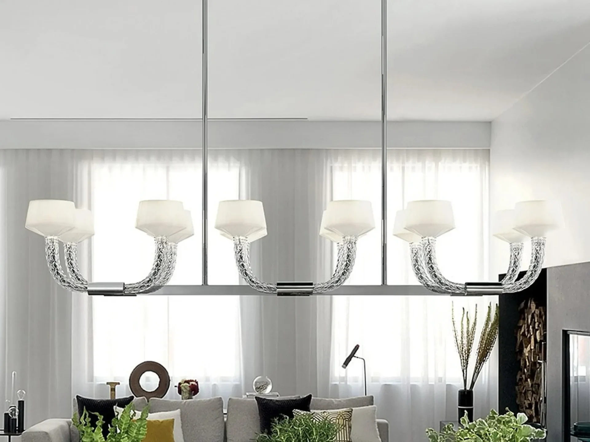 A modern chandelier with six white-shaded lights and clear, textured U-shaped arms, suspended from a white ceiling in a contemporary room with sheer white curtains.