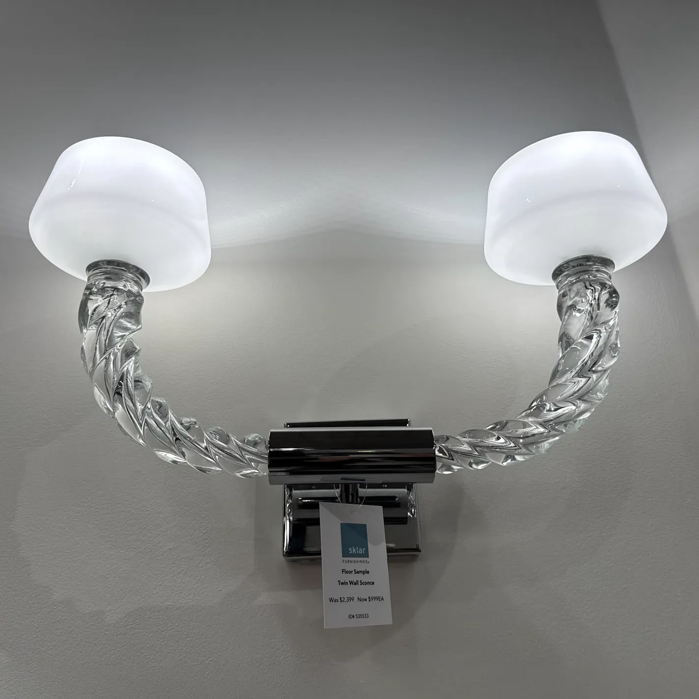 A twin wall sconce with two frosted glass shades and twisted clear glass arms, mounted on a light grey wall. A price tag for Sklar Furnishings is visible, indicating it's a floor sample on sale.