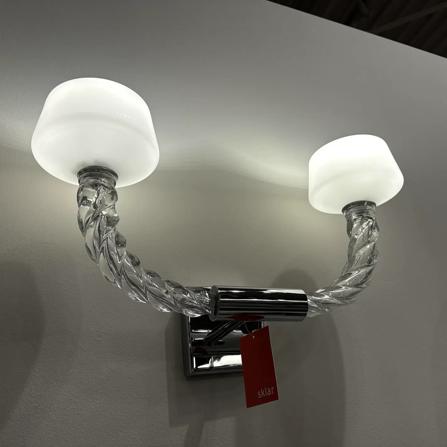 A two-light wall sconce with frosted white shades, clear twisted glass arms, and a chrome mounting plate on a light gray wall. A red 'sklar' tag hangs from the fixture.