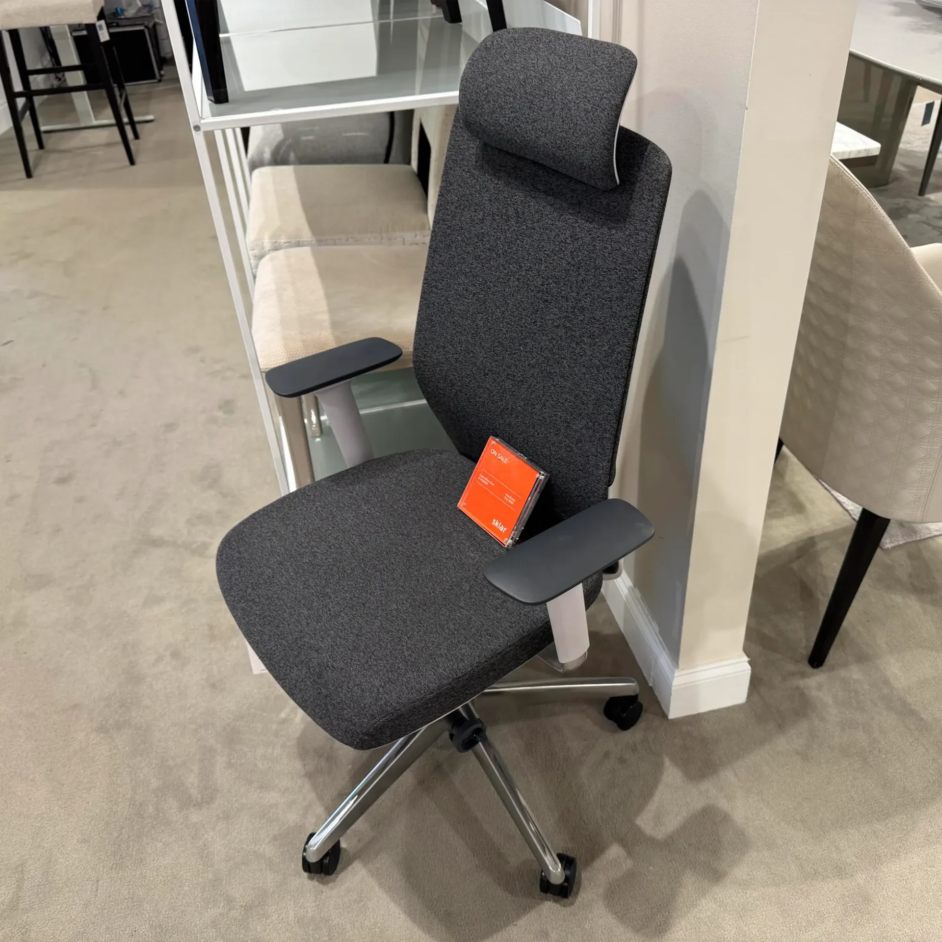 A dark gray ergonomic office chair with a high back and armrests, sitting on a carpeted floor with an orange 'ON SALE' sign on its seat.