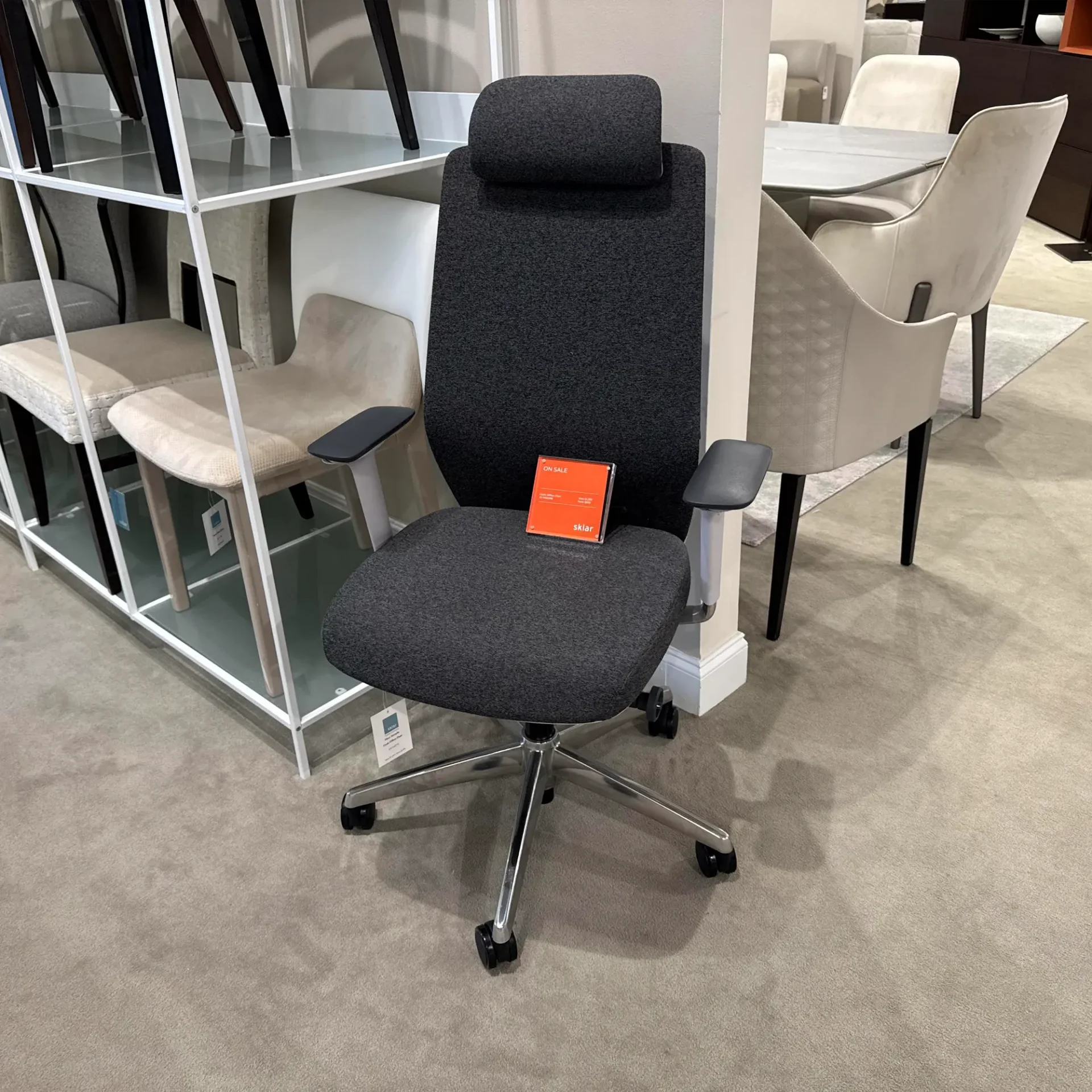 A dark gray ergonomic office chair with a headrest and armrests, featuring a chrome base and casters, sits on a gray carpet with an 'ON SALE' sign on its seat.