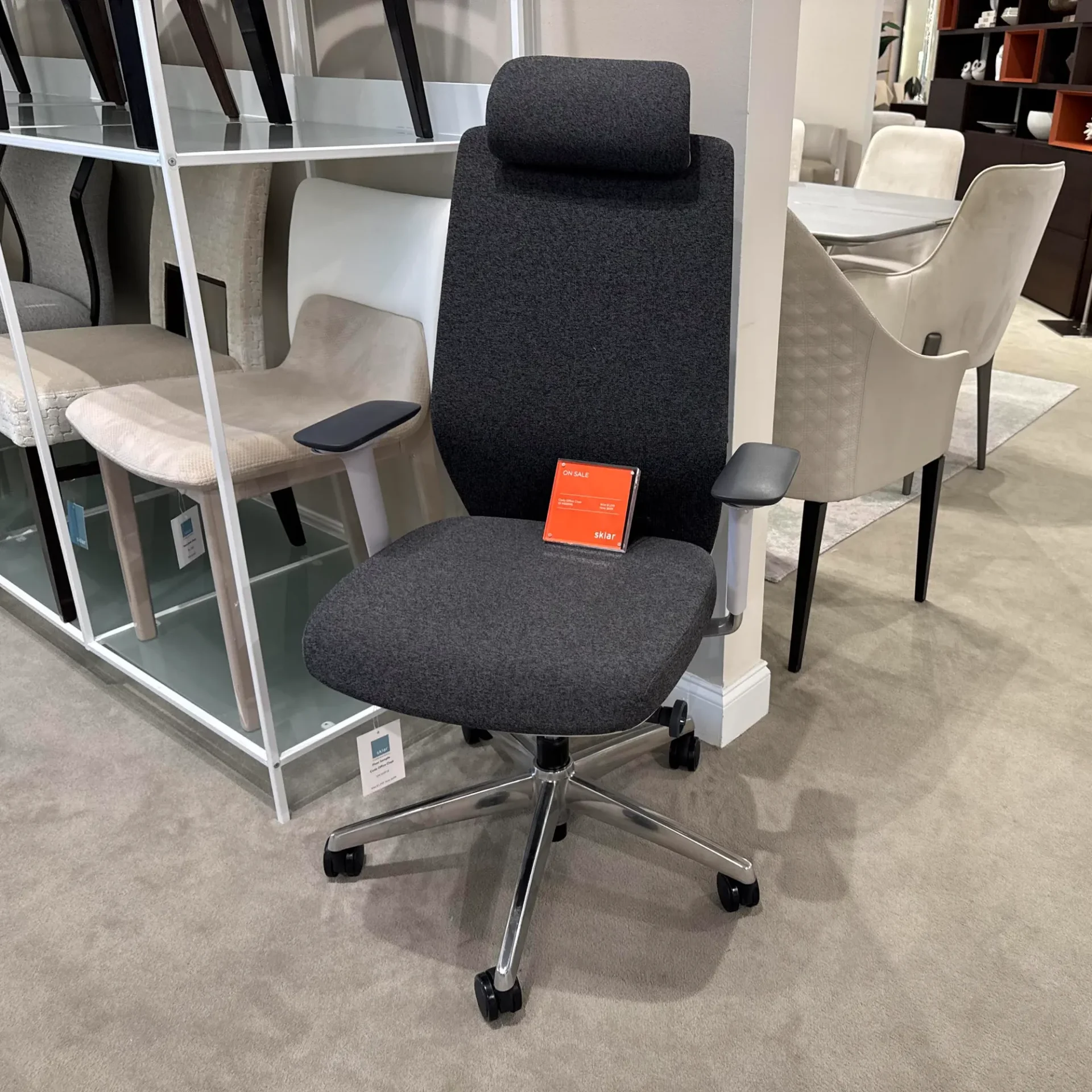 A dark gray office chair with a headrest, armrests, and wheeled base, featuring an orange 'ON SALE' sign on its seat, in a furniture showroom.