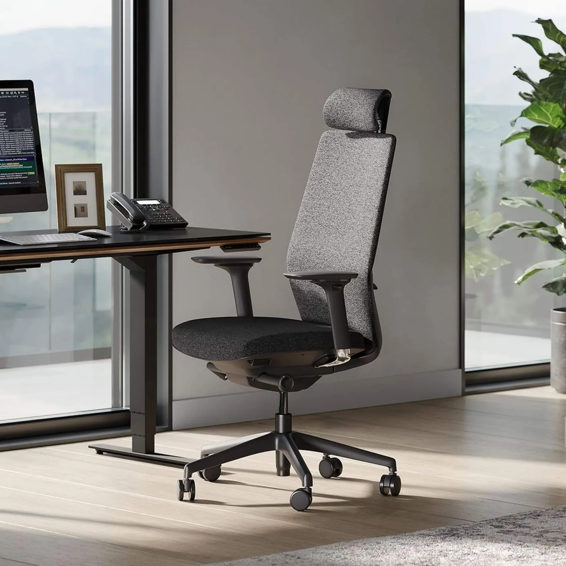 An ergonomic office chair with a gray back and black seat in front of a black standing desk with a computer, phone, and window view.