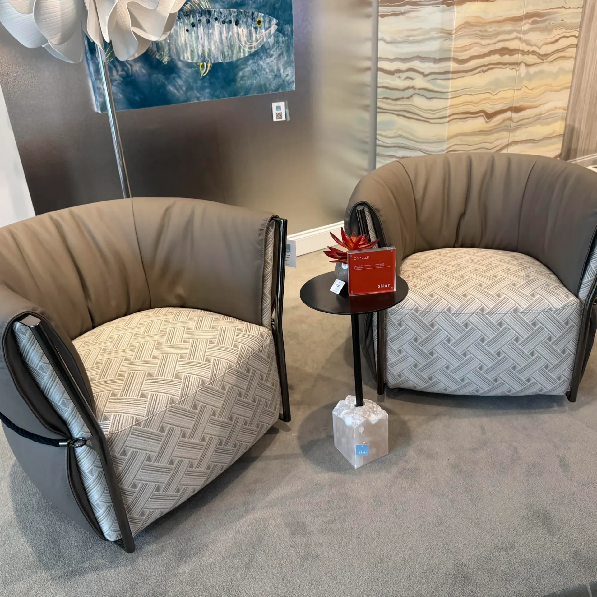 Two taupe and patterned fabric barrel armchairs flank a small black side table with a white stone base. A red 'ON SALE' sign and a succulent are on the table. A fish artwork hangs on the wall behind.