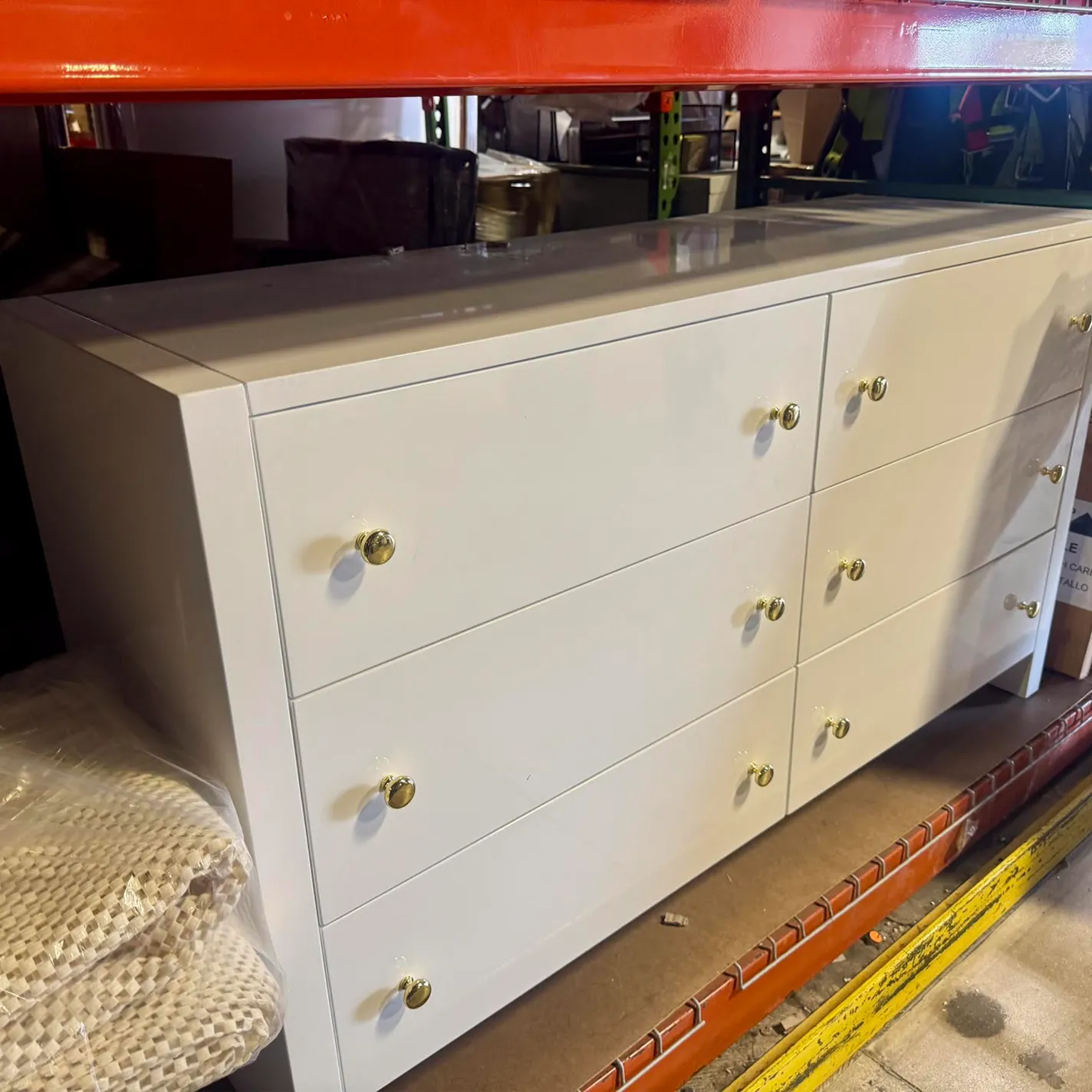 A white, glossy dresser with six drawers and gold knobs, seen in a storage area under an orange shelf.