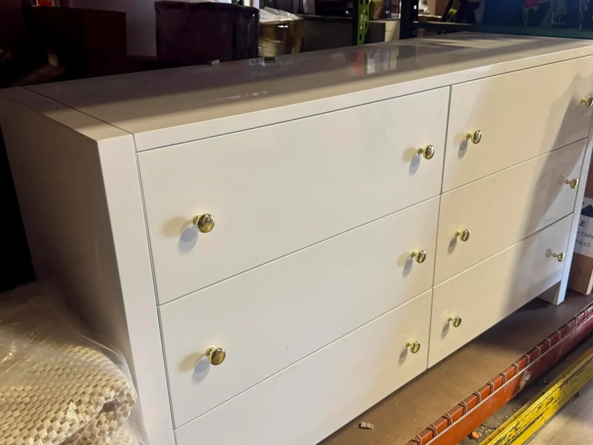 A white, glossy six-drawer dresser with gold knobs in a storage area.