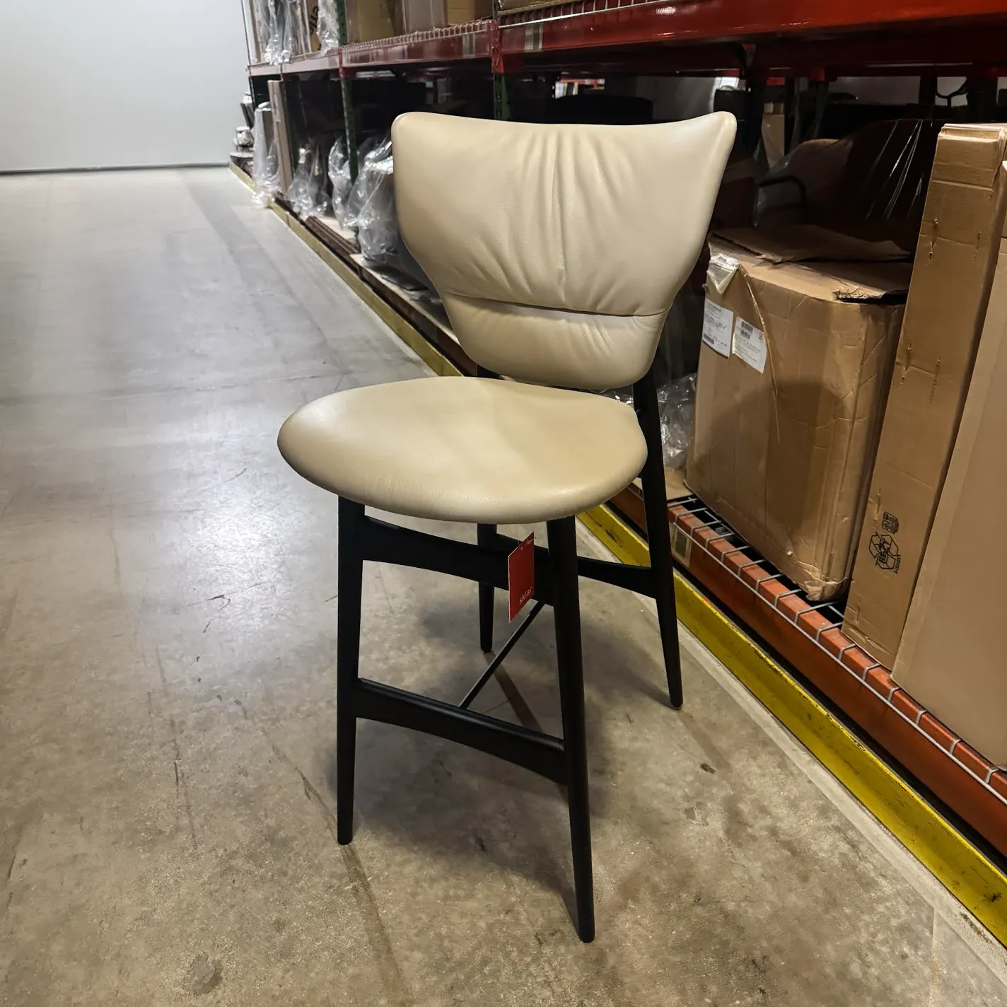 A beige upholstered chair with a black frame sits on a concrete floor in a warehouse, surrounded by shelves and boxes.