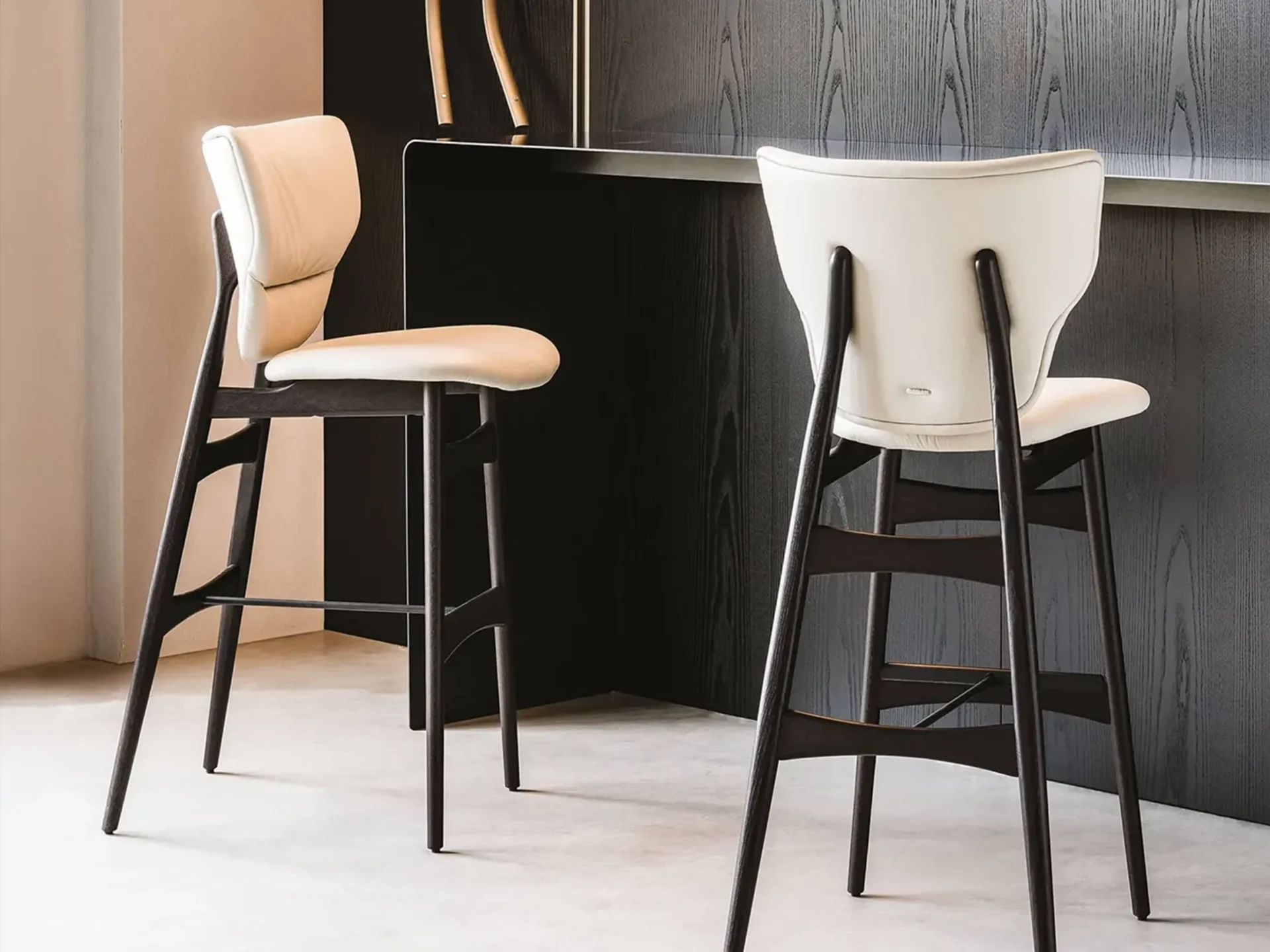 Two modern bar stools with cream upholstery and dark wood frames, set against a dark wood paneling and light wall.