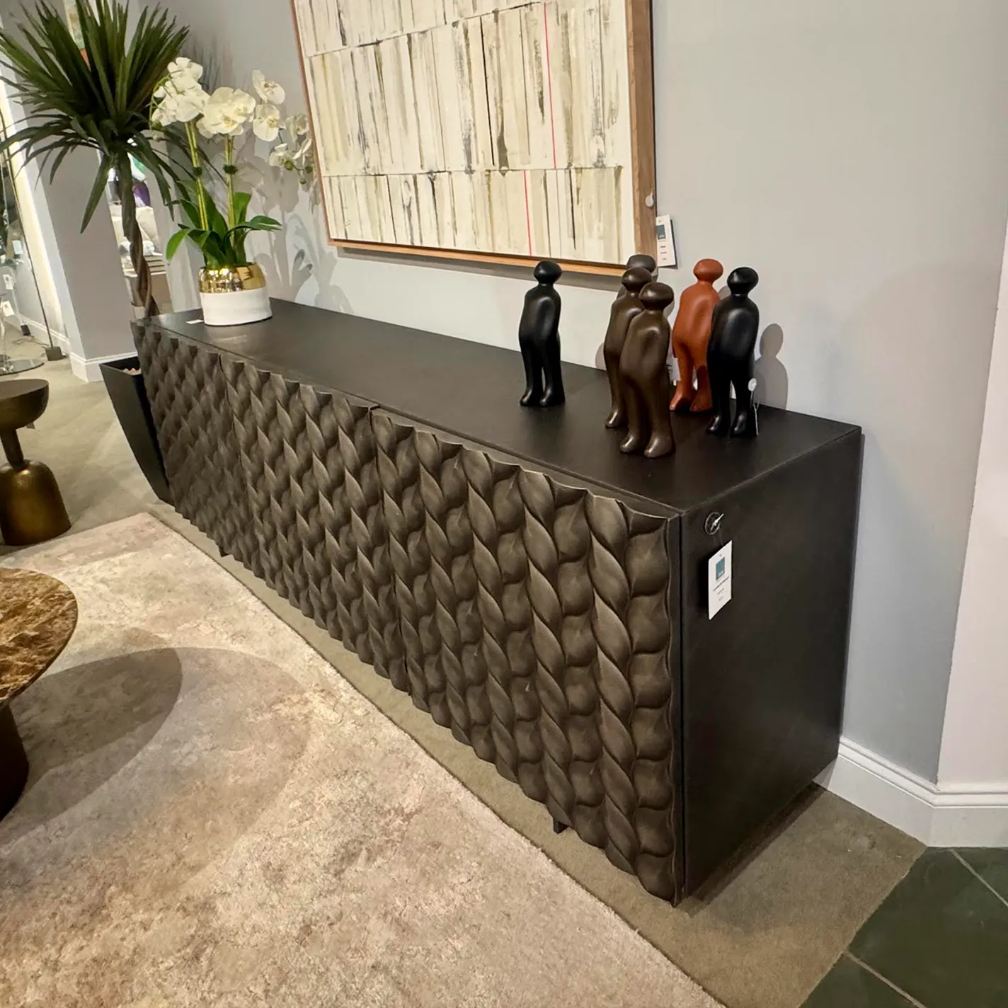 A dark gray console table with a textured, woven-like front, topped with white orchids and five stylized humanoid figurines, set against a light gray wall with abstract art.