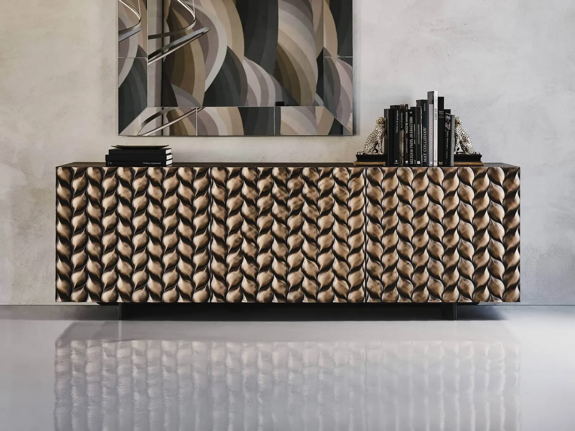 A modern living room featuring a long, low sideboard with a textured, bronze-colored, leaf-patterned front. On top are books and leopard figurines, with a segmented mirror reflecting abstract art above, all set against a gray wall and reflective floor.
