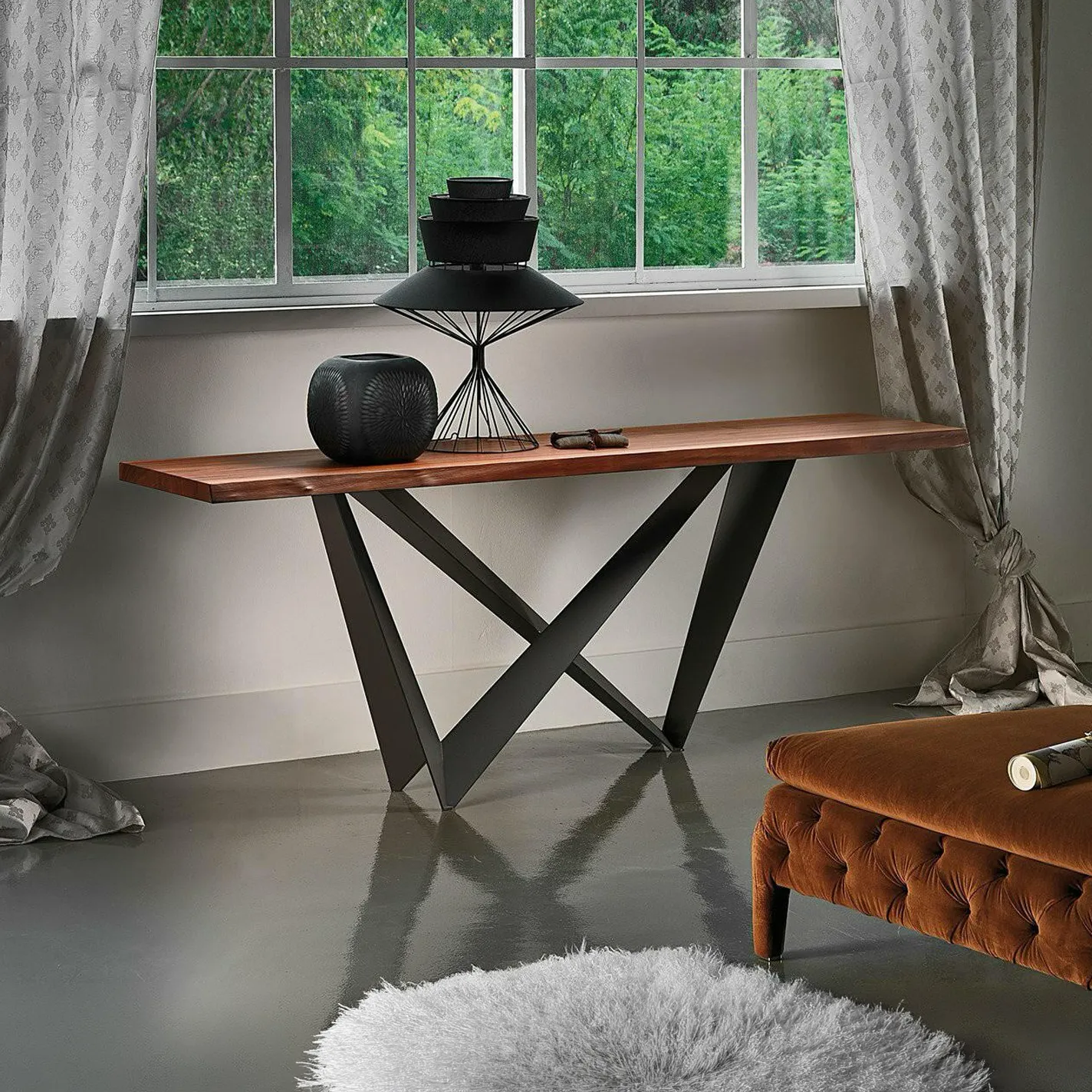 A modern console table with a wooden top and black metal legs, adorned with black decorative objects, sits in front of a window with green foliage outside. A fluffy rug and a brown tufted ottoman are also visible.