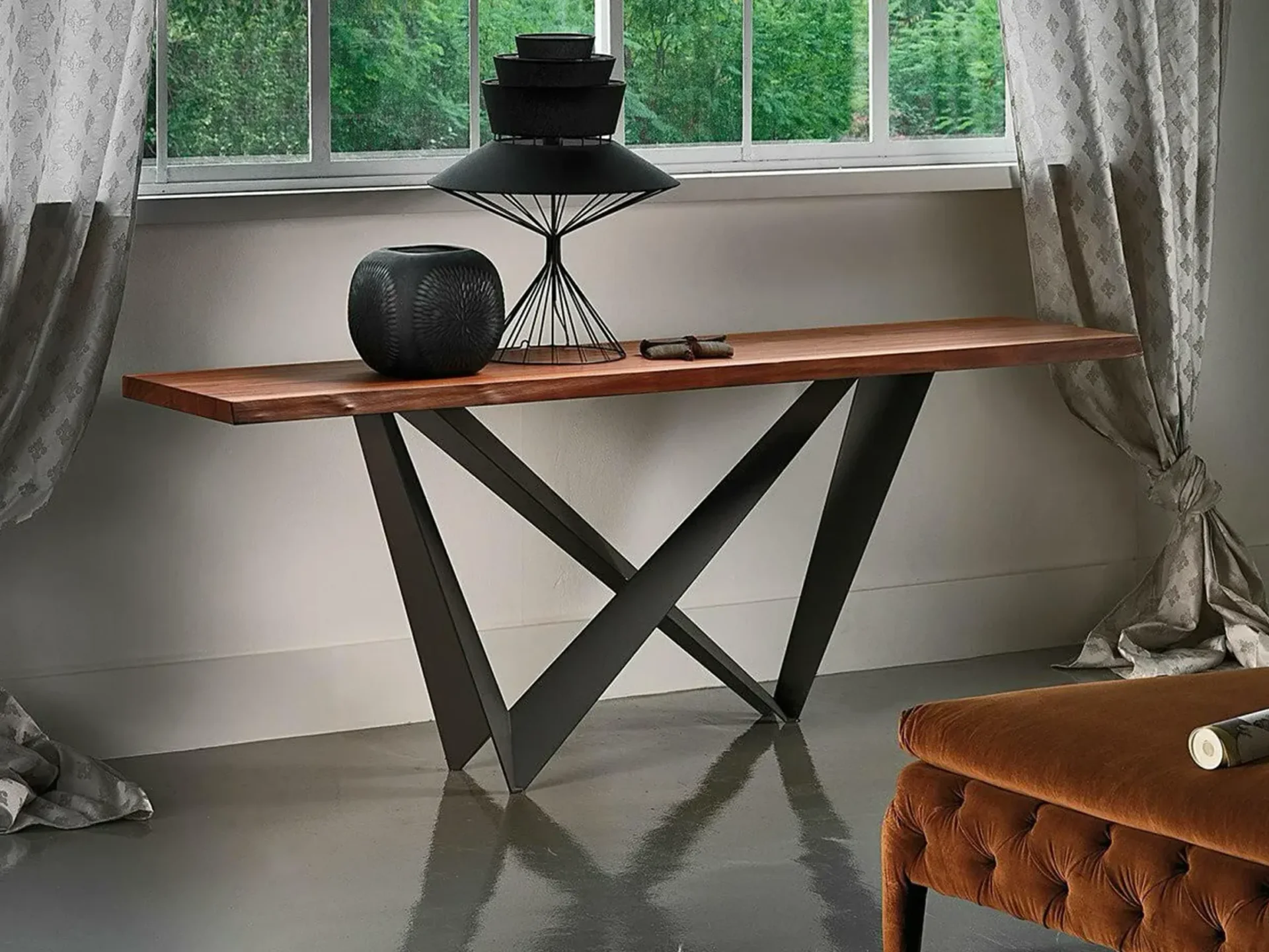 A modern console table with a wooden top and black geometric metal base, adorned with black decorative objects, in front of a window with green foliage and gray curtains.