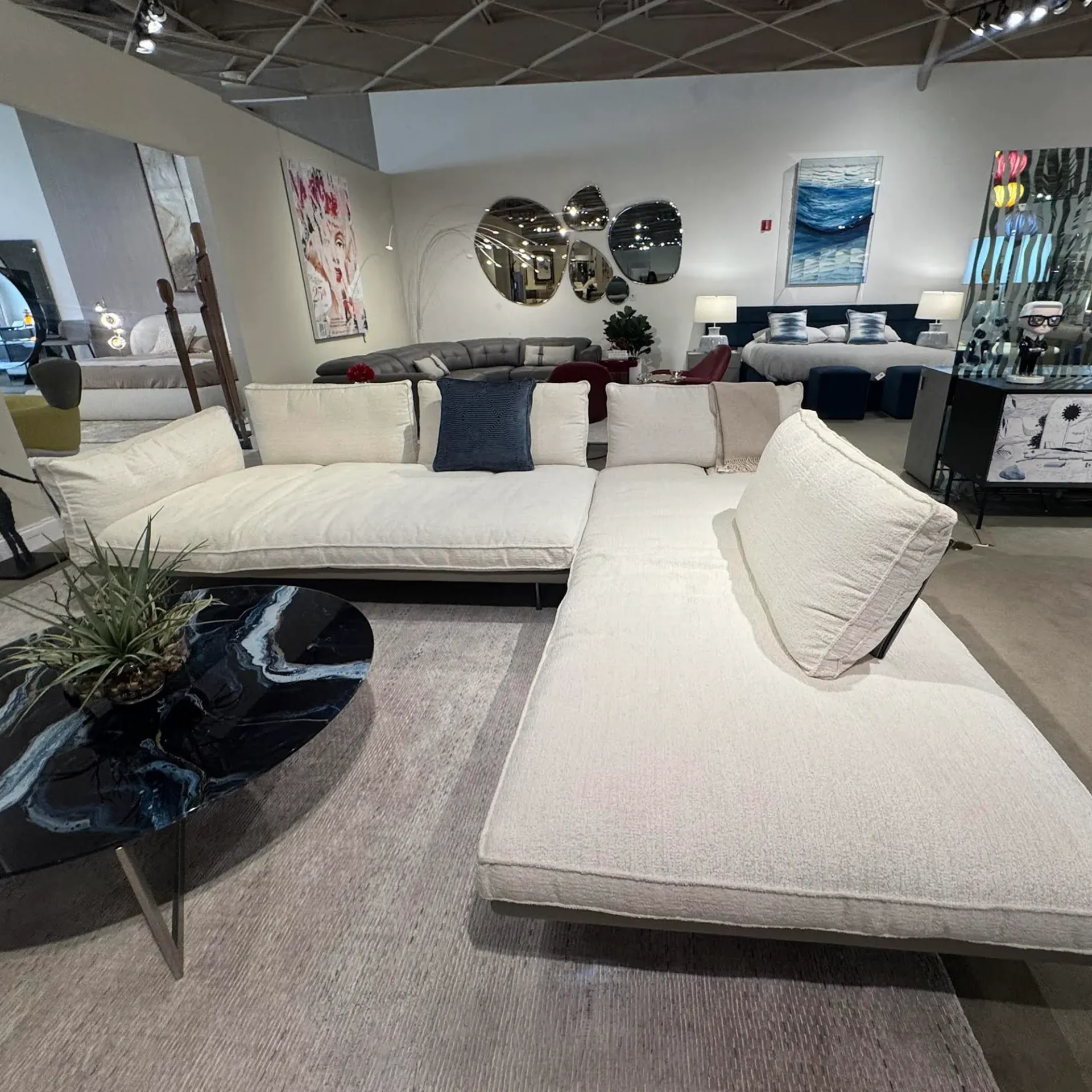 A large, L-shaped white sectional sofa with a dark blue throw pillow and a black marbled coffee table with a plant, set on a gray rug in a furniture showroom.