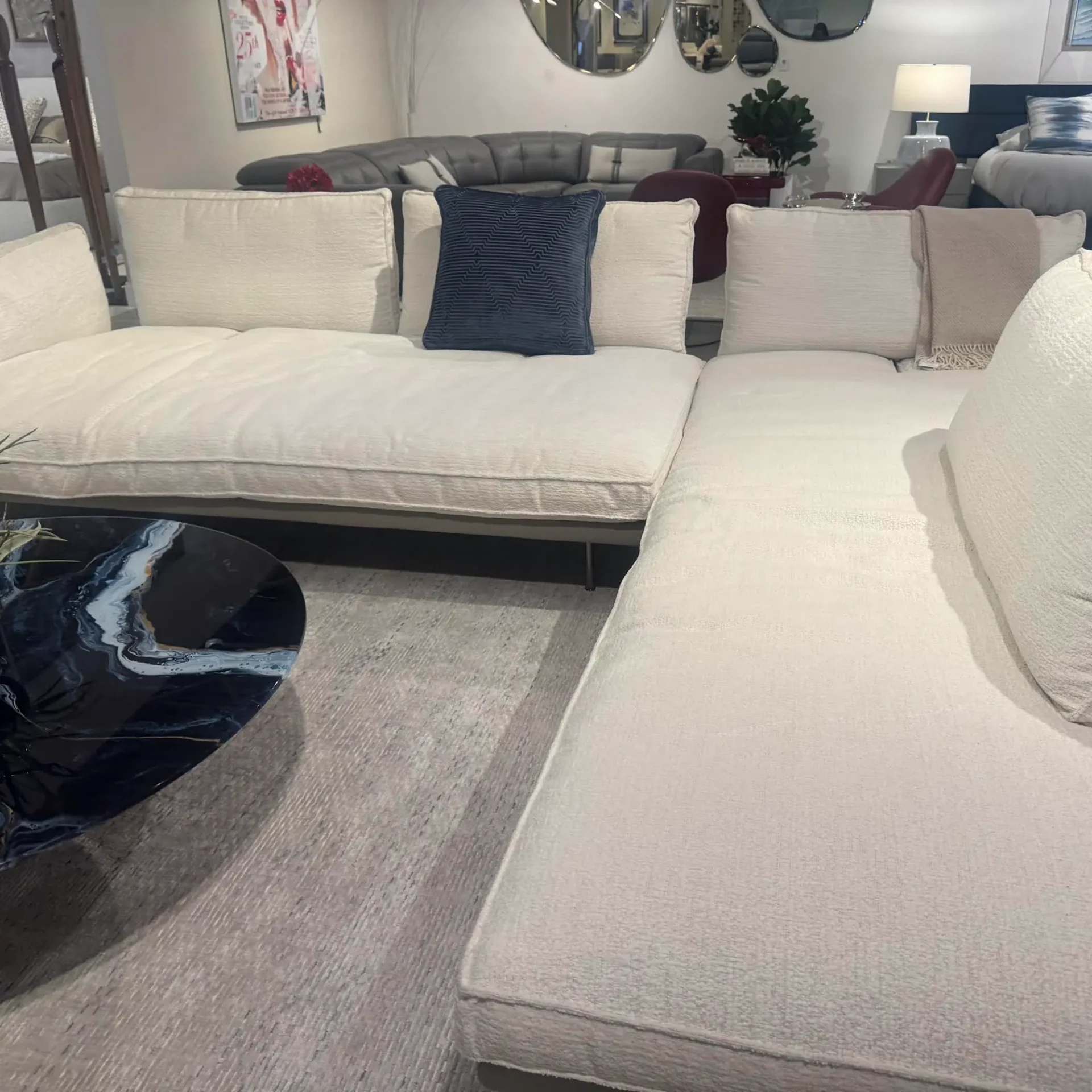 A large, L-shaped off-white sectional sofa with a navy blue pillow and a beige throw blanket. In front is a round, dark coffee table with a blue and white abstract pattern. A gray rug is on the floor, and other furniture is visible in the background.