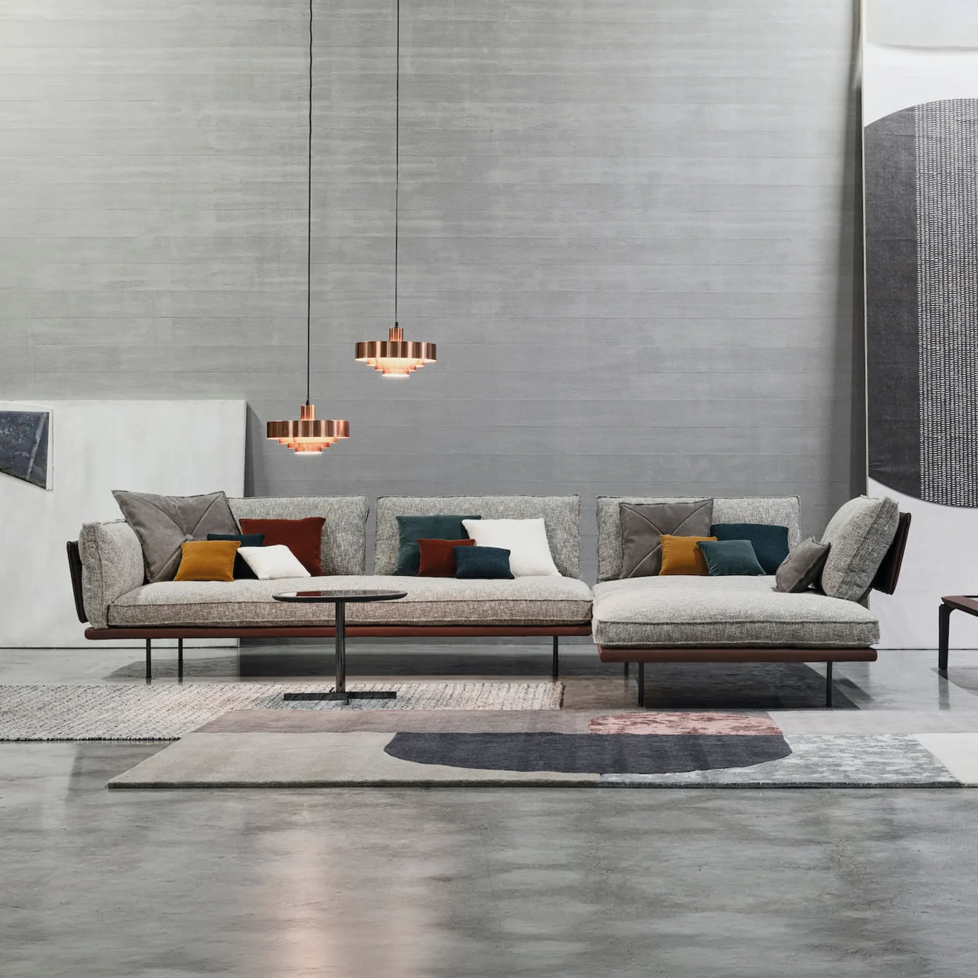 An L-shaped sectional sofa with light grey fabric and colorful pillows, a round coffee table, and layered rugs on a concrete floor, under two copper pendant lights.