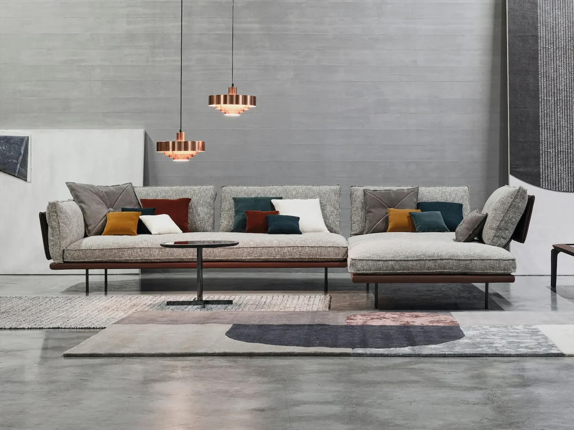 A modern living room with a large grey sectional sofa, colorful throw pillows, a black coffee table, layered rugs, and copper pendant lights against a concrete wall.