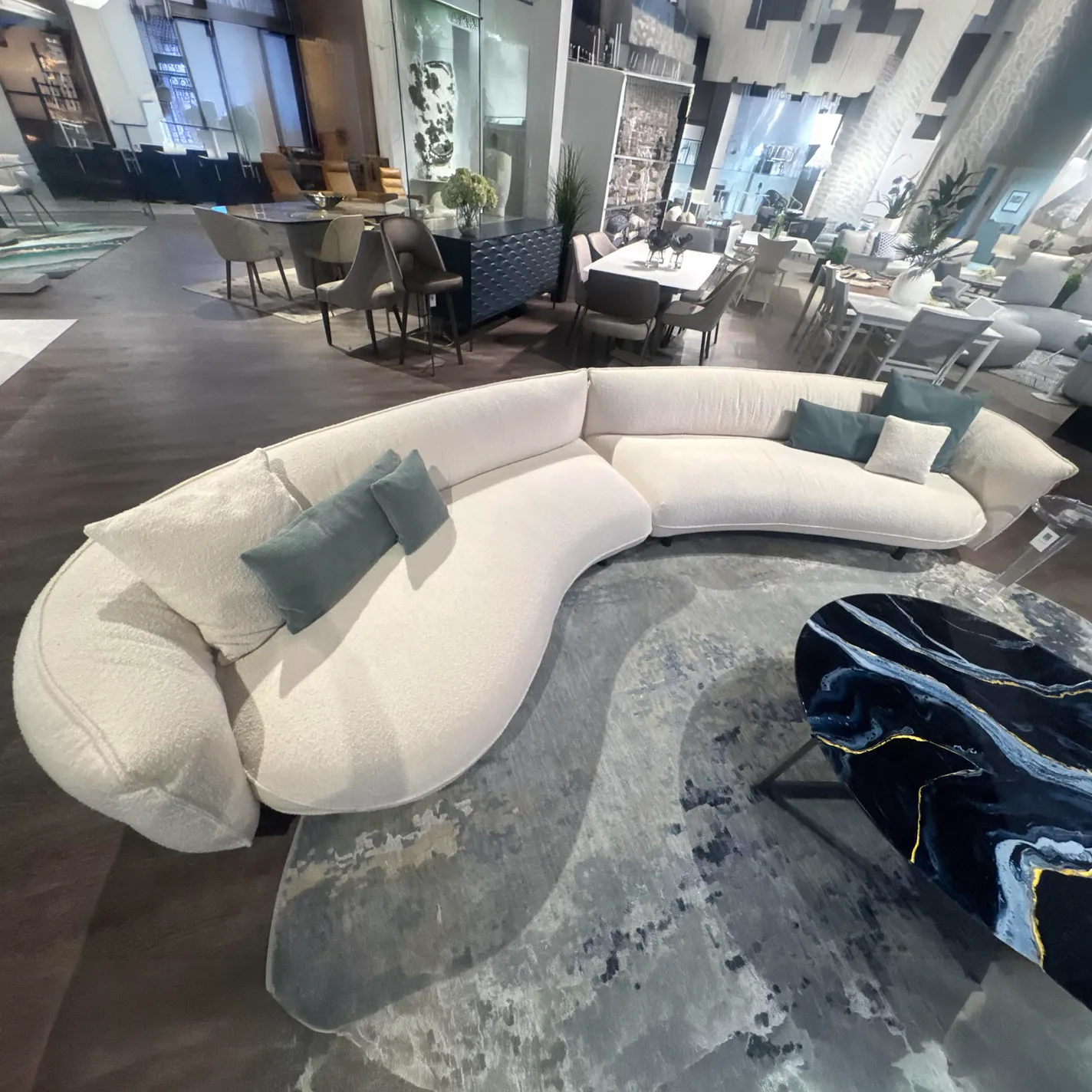 A large, curved off-white modular sofa with teal pillows on an abstract grey rug, next to a dark blue and gold marble coffee table, in a modern furniture showroom.