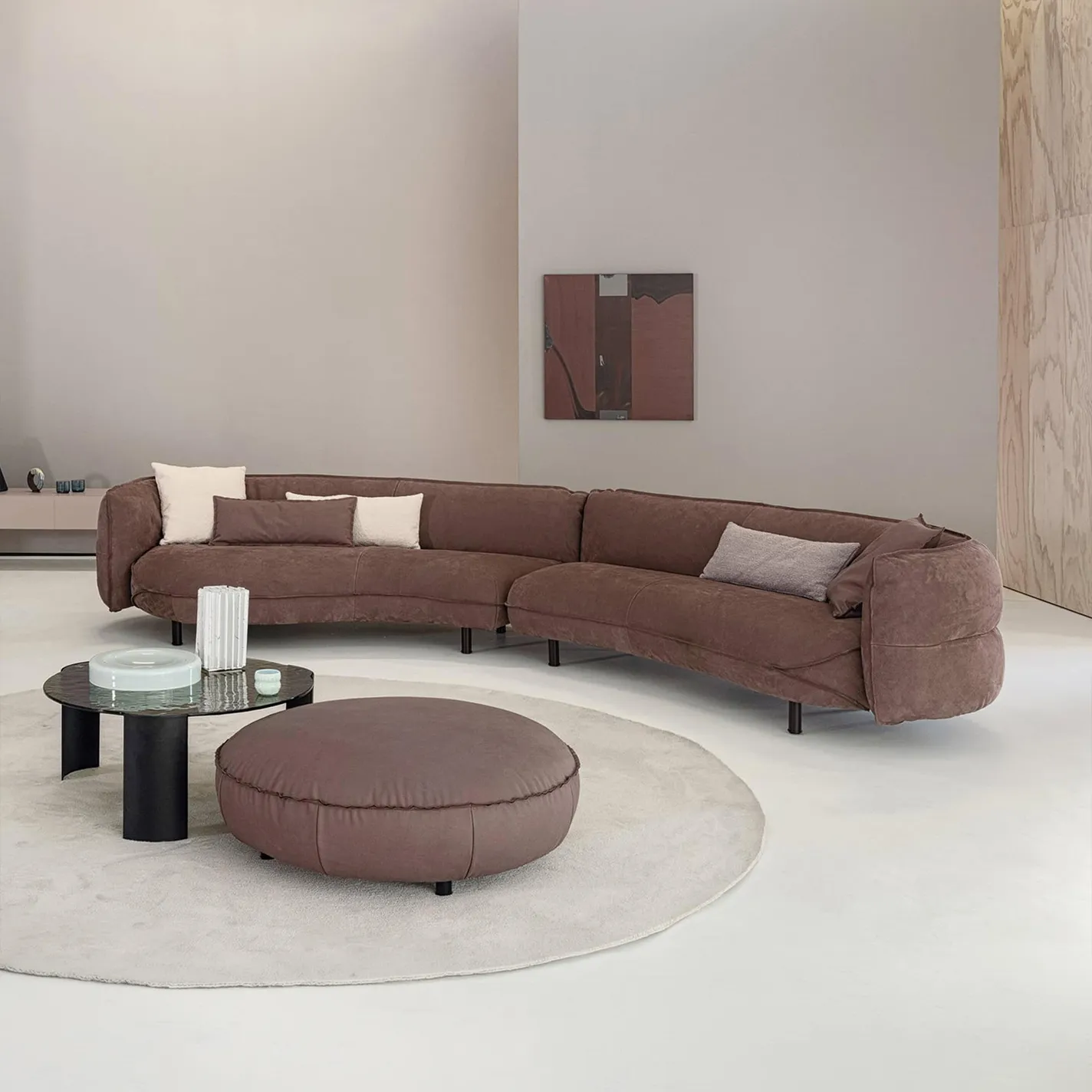 A modern living room with a large, curved brown sectional sofa, a round coffee table with glass decor, and a matching brown ottoman on a cream rug. The walls are neutral with an abstract painting and wood paneling.