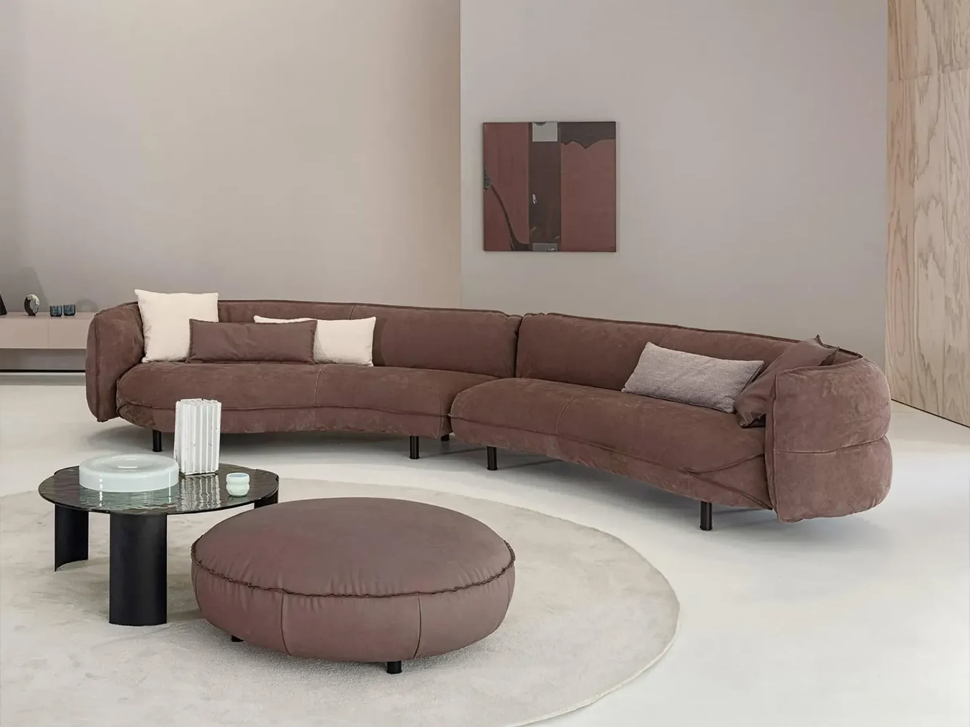 A modern living room featuring a large, curved brown sectional sofa with throw pillows, a round glass coffee table, and a matching brown ottoman on a light rug. An abstract painting hangs on the neutral wall, and a wood-paneled wall is visible on the right.
