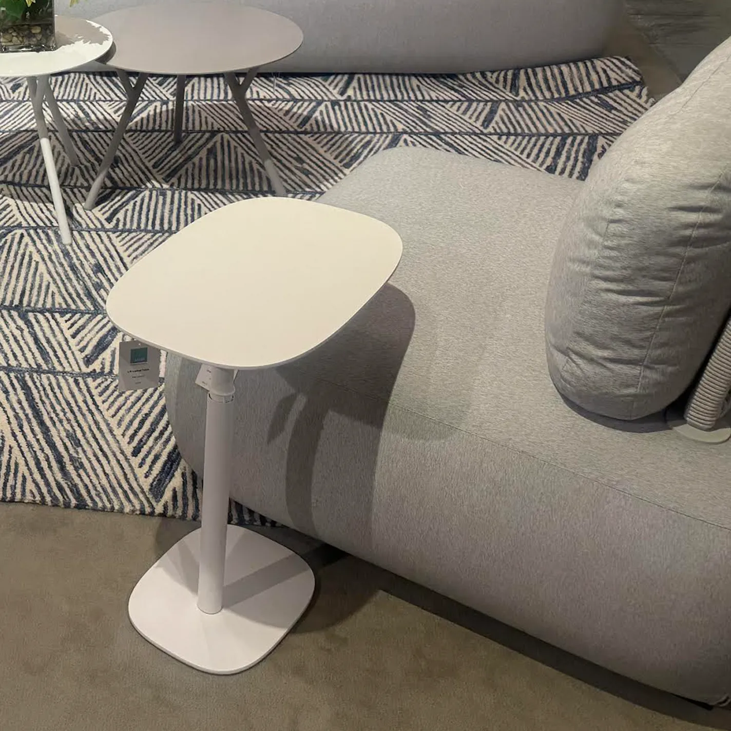 A white adjustable side table with a rounded square top next to a light gray sofa, on a blue and white geometric rug. Other tables are in the background.