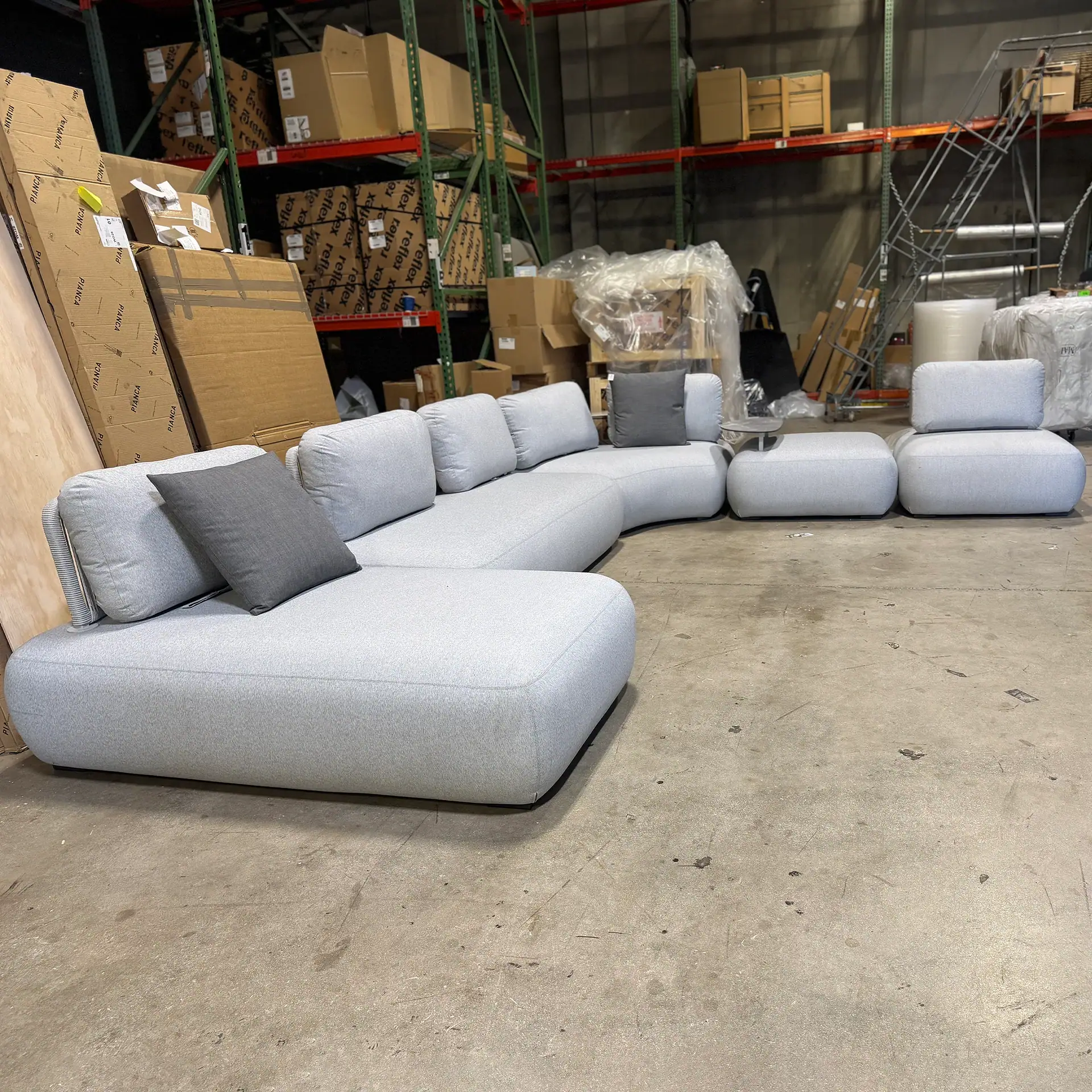 Modern outdoor sectional sofa with light gray cushions and pillows in warehouse setting.