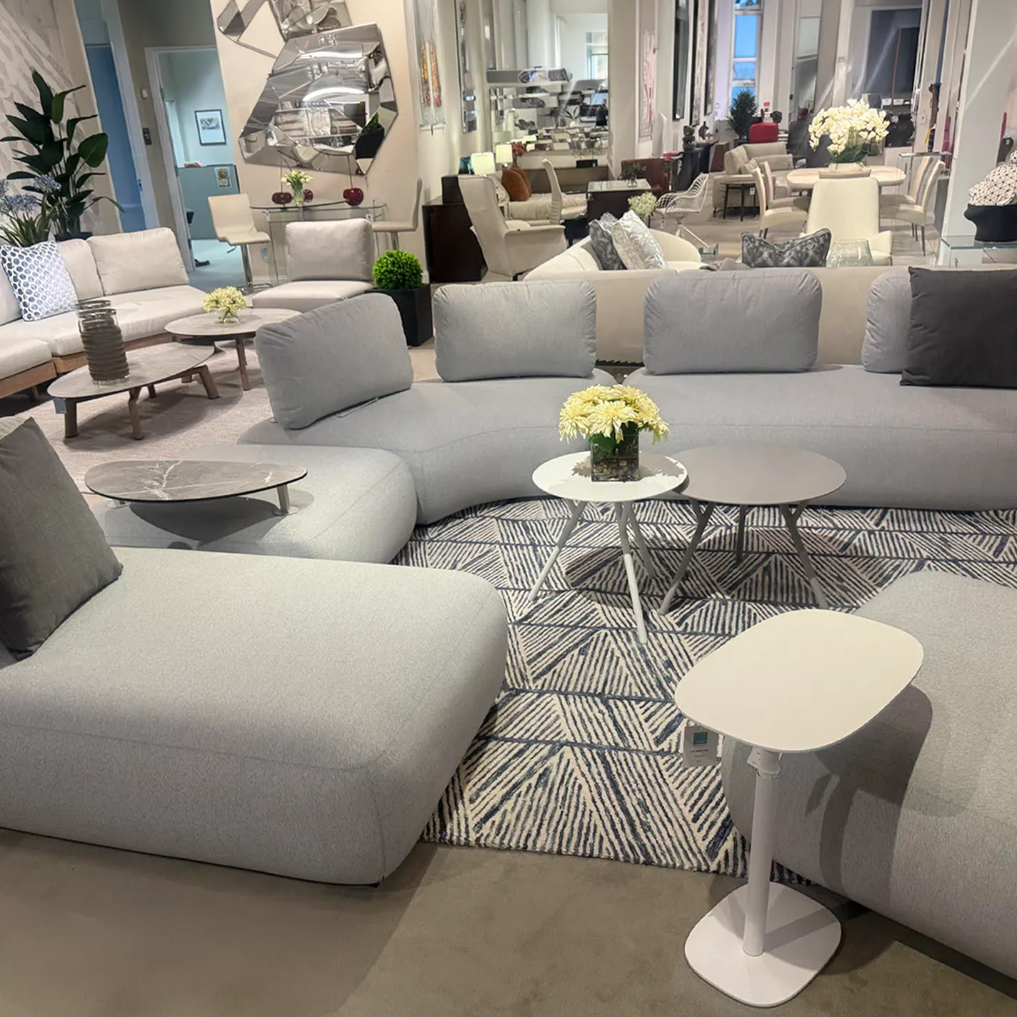 A large, light gray modular sofa with rounded edges in a modern furniture showroom, surrounded by various coffee tables and a geometric patterned rug.
