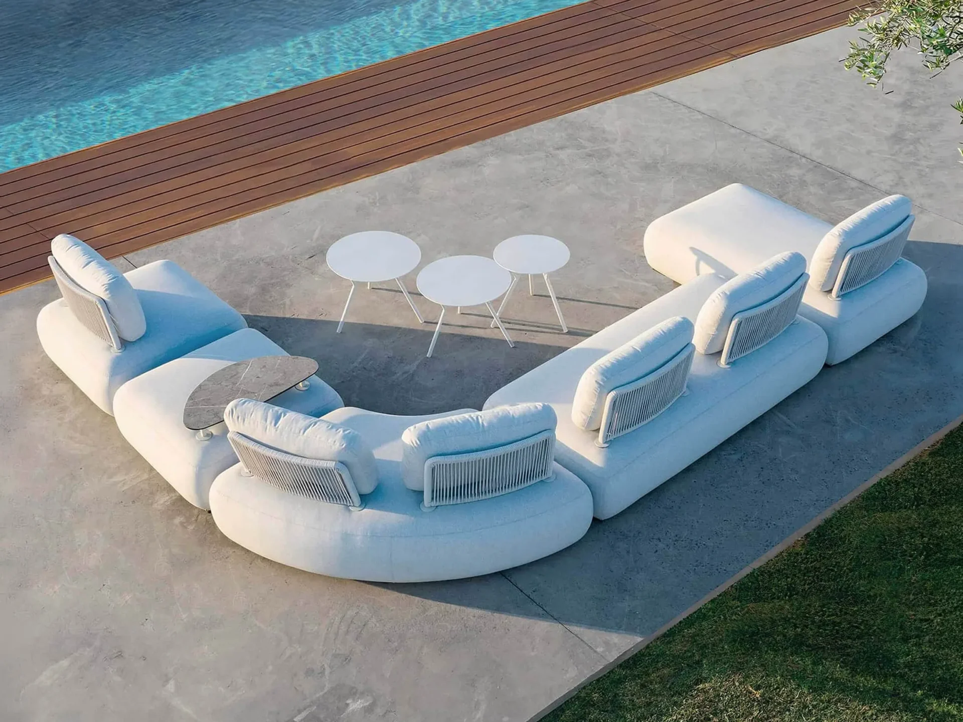 Aerial view of a white modular outdoor sofa with integrated tables on a concrete patio next to a swimming pool with a wooden deck and green grass.