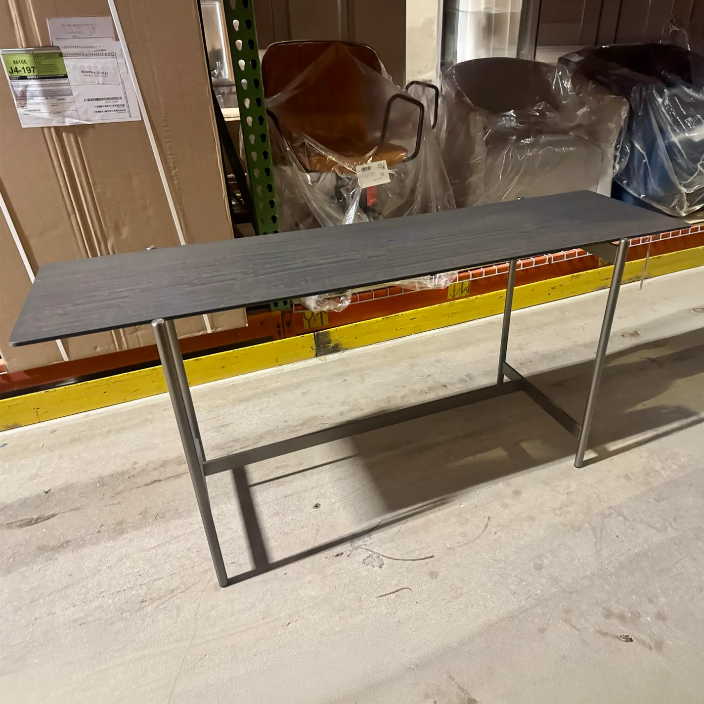 A dark gray console table with metal legs in a warehouse. Cardboard boxes and plastic-covered furniture are in the background.