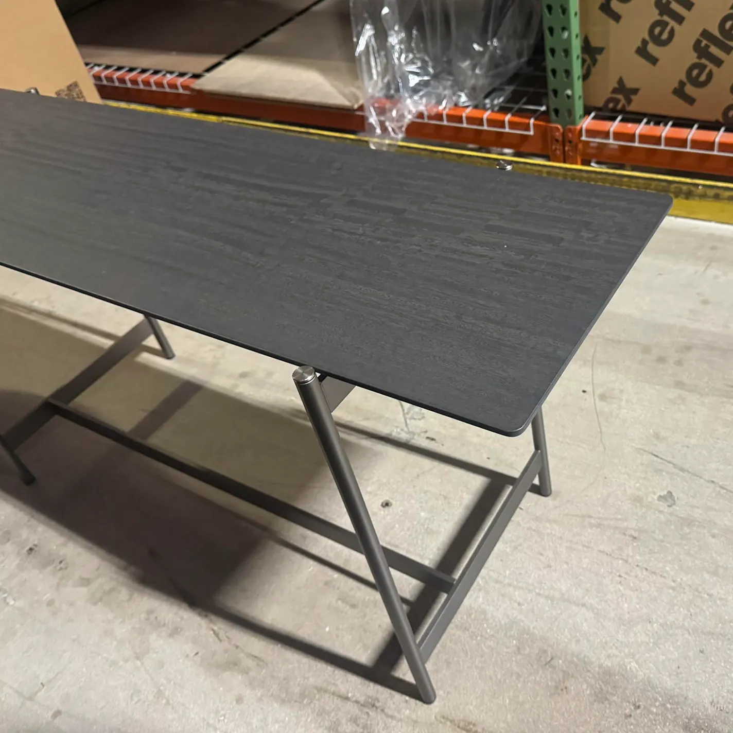 A dark gray rectangular table with textured surface and angled gray metal legs on a concrete floor, with warehouse shelving in the background.